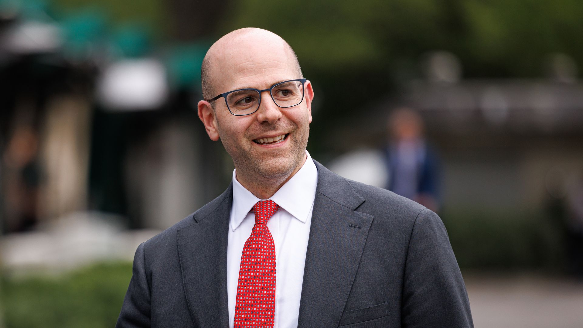 Smiling man with glasses wearing a dark suit, white shirt, and red patterned tie, outdoors with blurred greenery in the background.