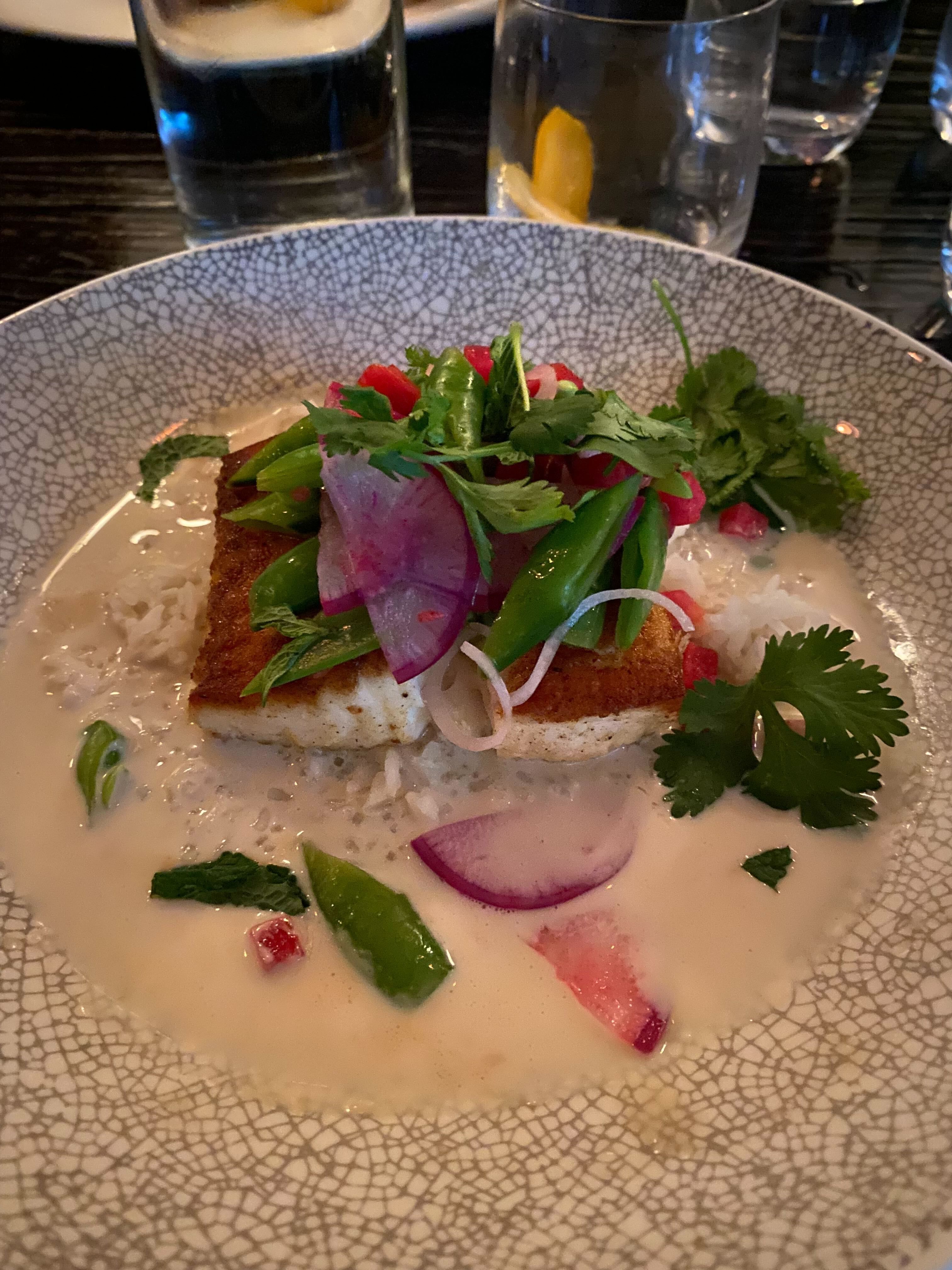 Plate with a browned fish fillet topped with green snap peas, purple radish slices, and cilantro, served over white rice and creamy light-colored sauce, on a textured plate.
