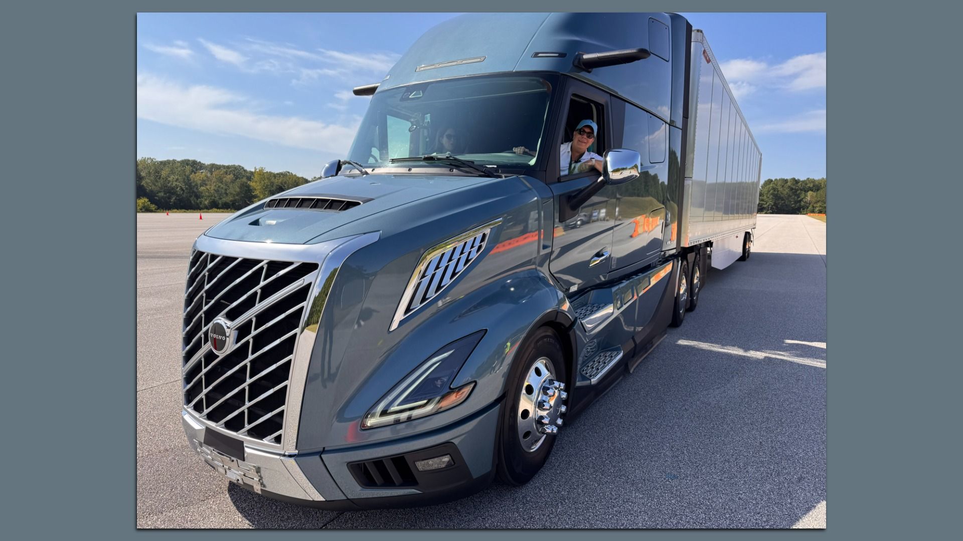 Author Joann Muller leans out the window of a blue Volvo semi truck, on a paved open road on a sunny day.