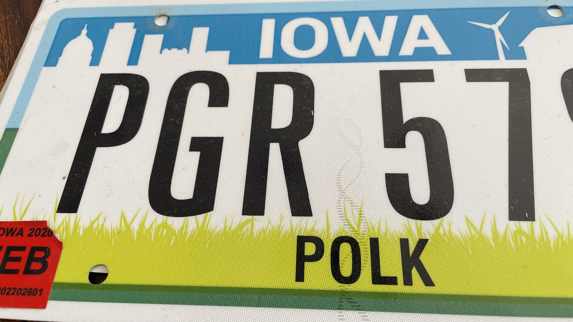 Close-up of an Iowa license plate: blue header with IOWA and skyline, large black characters PGR 5 1 across the center, POLK county text at the bottom, a yellow grass motif along the edge, and a red validation tag in the lower left.