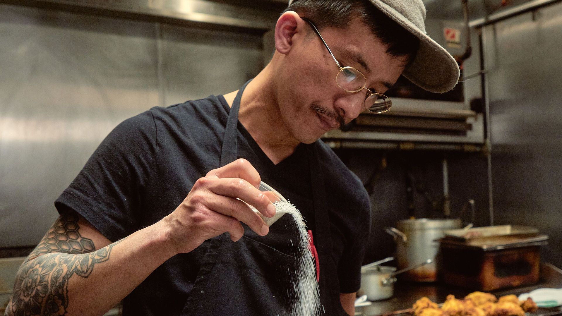 Chef Ethan Lim wears a black apron in the kitchen and pours salt from a small cup.