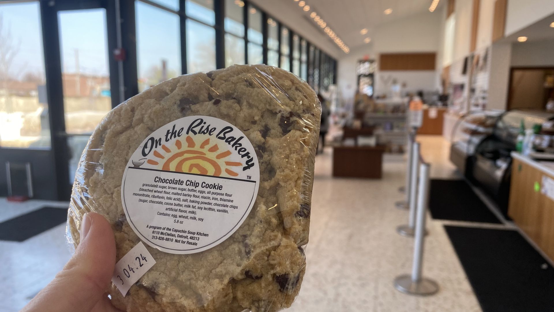 A cookie held aloft in front of a sunny bakery with lots of windows.