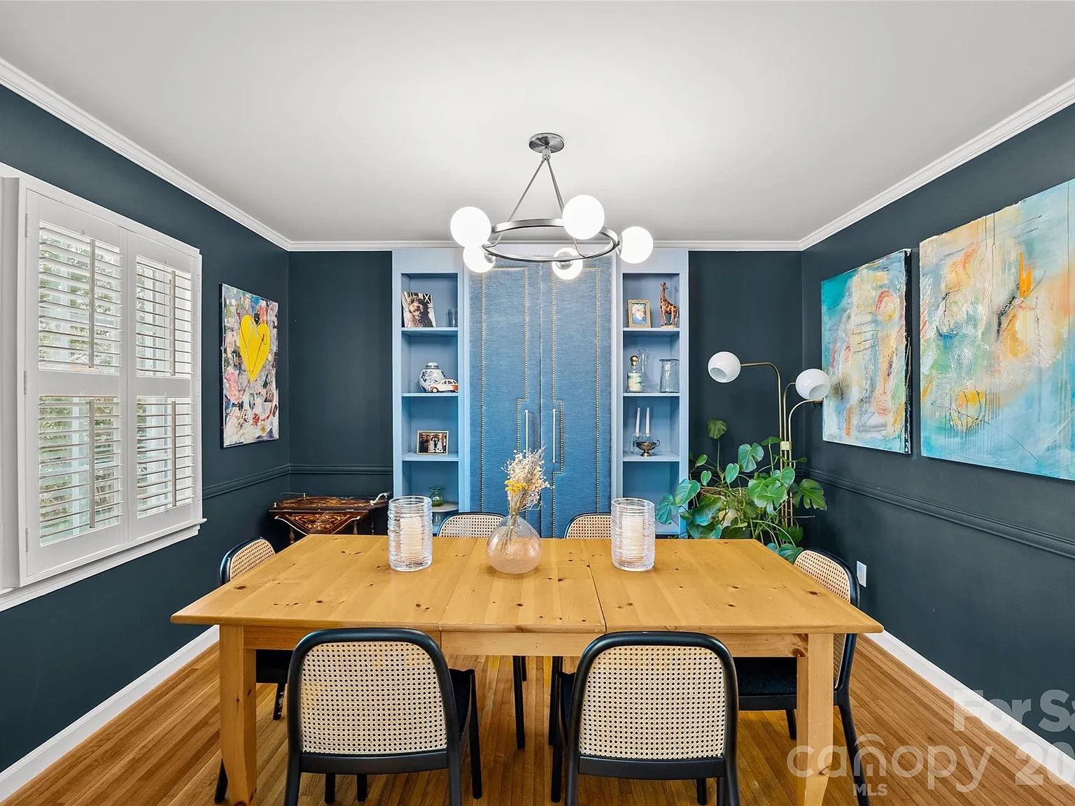 Dining room with dark blue walls, light wood table, and cane chairs. A chandelier hangs above a blue built-in cabinet; abstract art and a yellow heart painting adorn the walls, with window and plant.