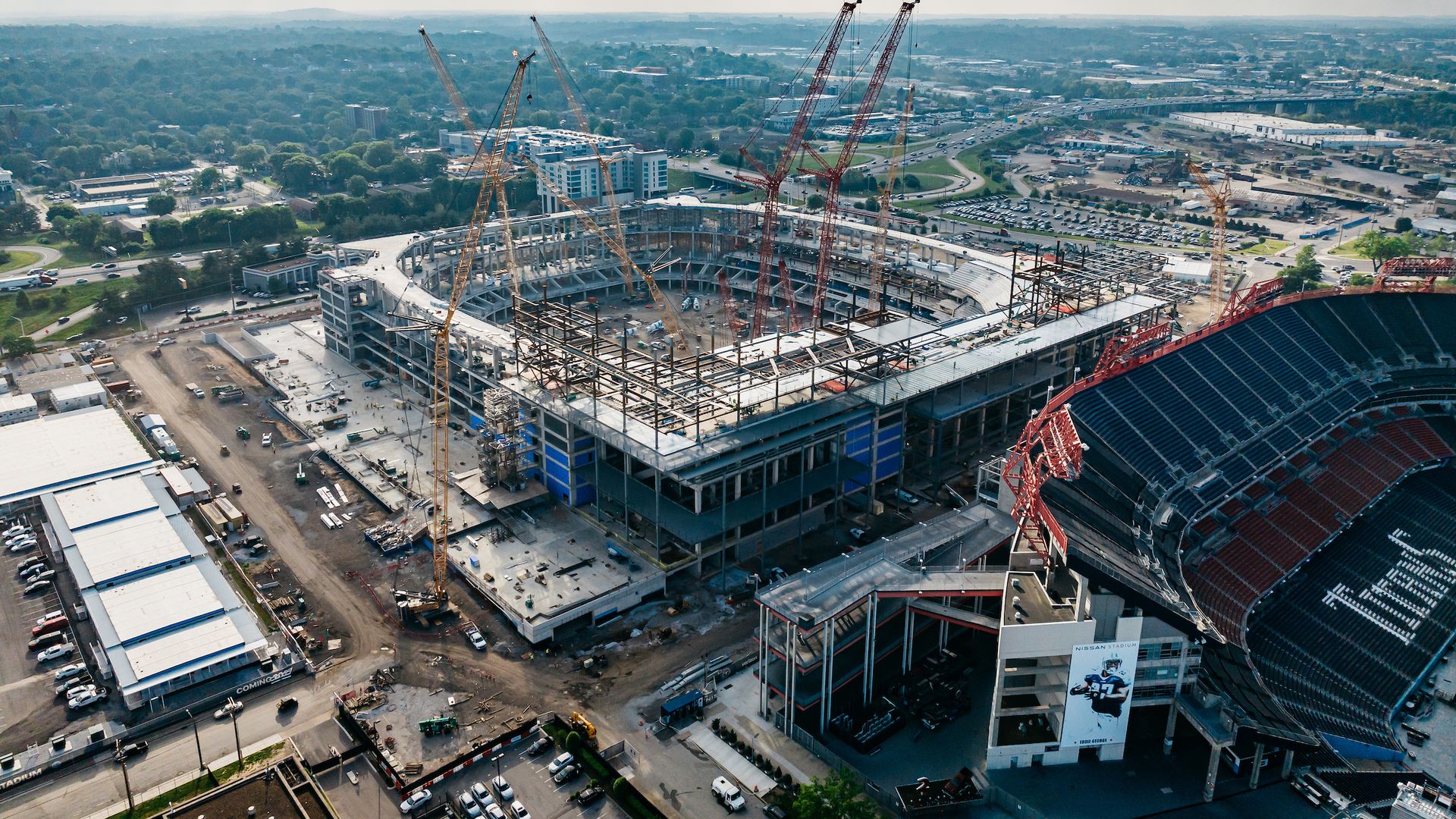 A large stadium under construction with cranes around it next to a completed stadium section with red and black seats and the name Nissan Stadium visible, under a cloudy sky.