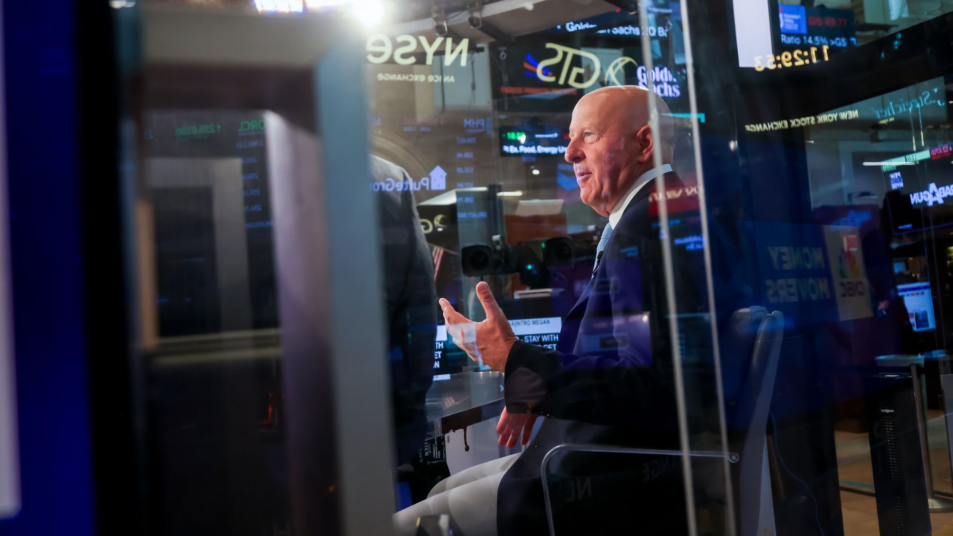 Man in business suit speaking inside a glass-enclosed studio with stock market ticker displays and financial logos like NYSE and Goldman Sachs in the background.