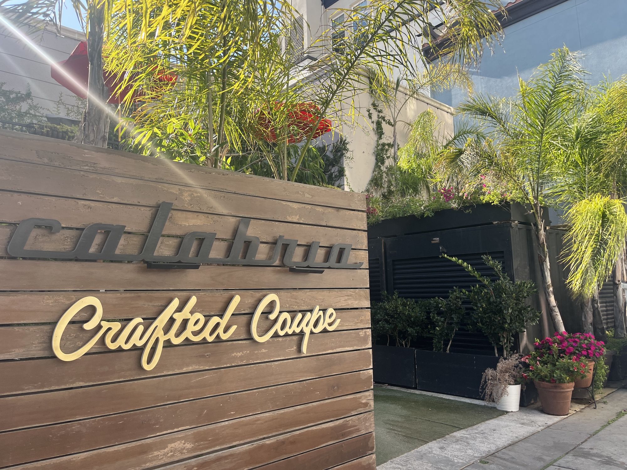 Wooden sign with black and gold text "calabria Crafted Coupe" surrounded by palm trees and potted plants in a sunny outdoor setting.