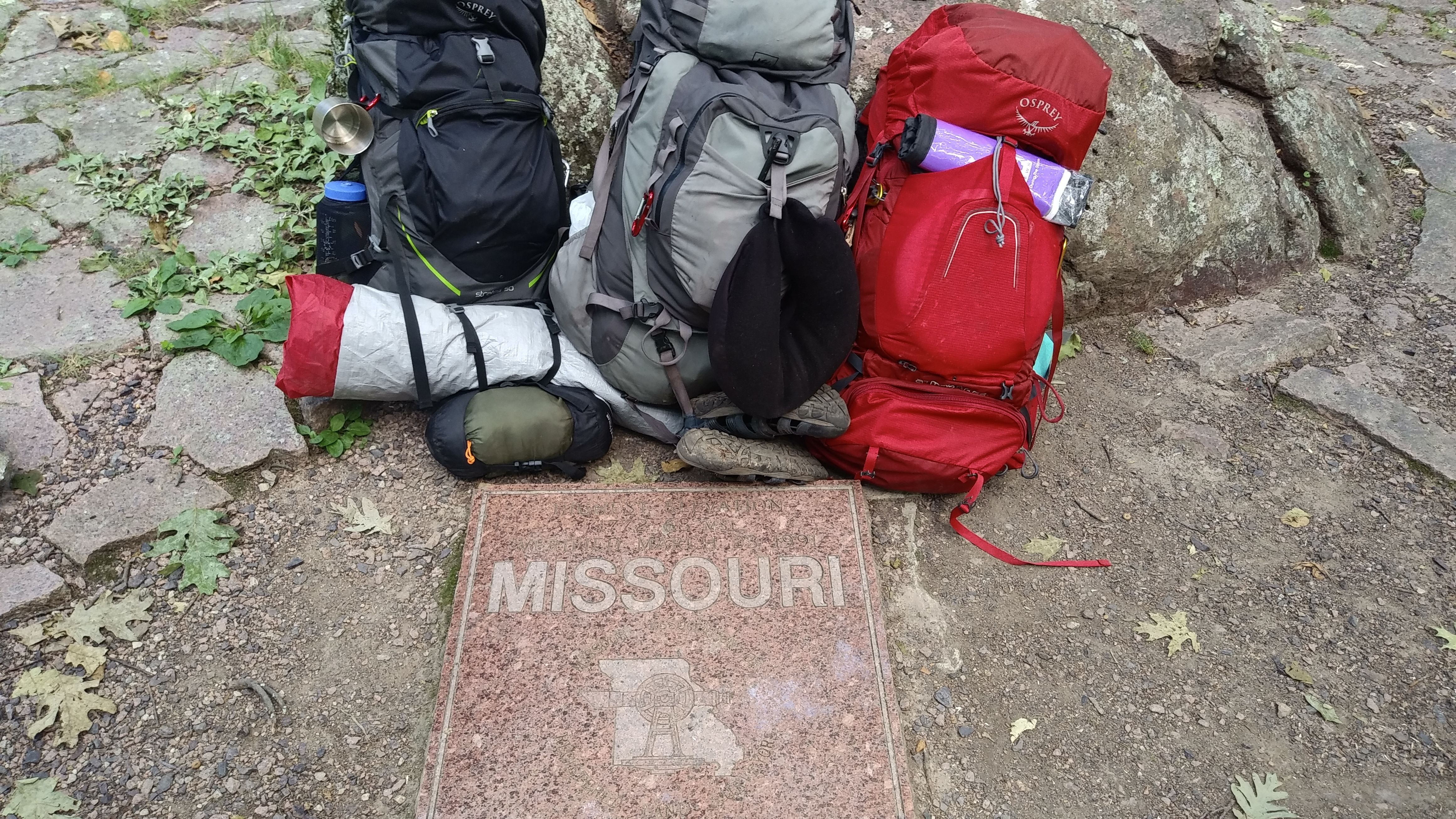 Three loaded hiking backpacks (black, gray, red) rest against a rocky wall beside a Missouri marker plaque. Gear includes a rolled mat, water bottle, metal cup, and hiking boots.