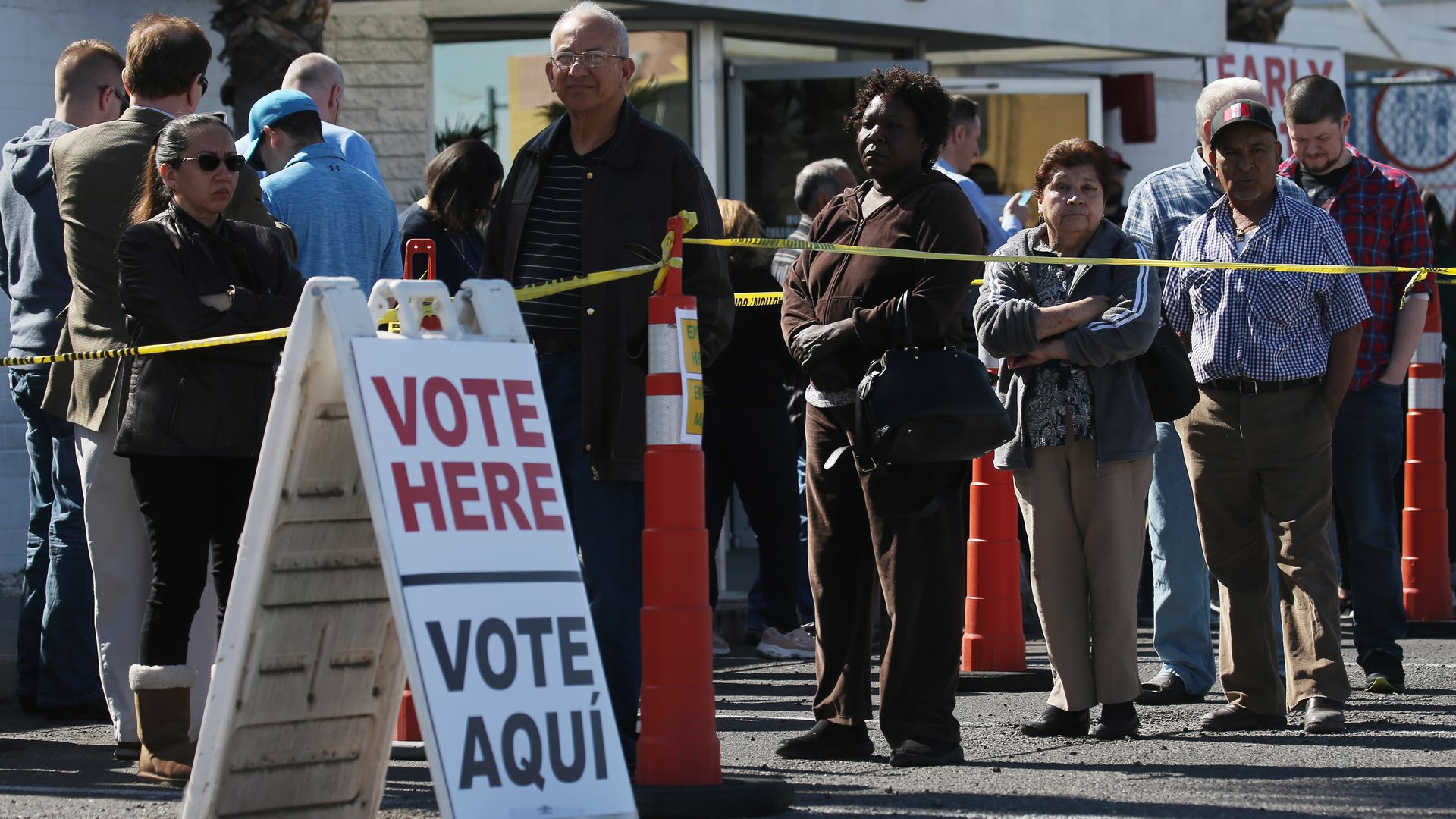 In this image, a line of people wait outside near a sign that reads "vote here" and vote aqui" 