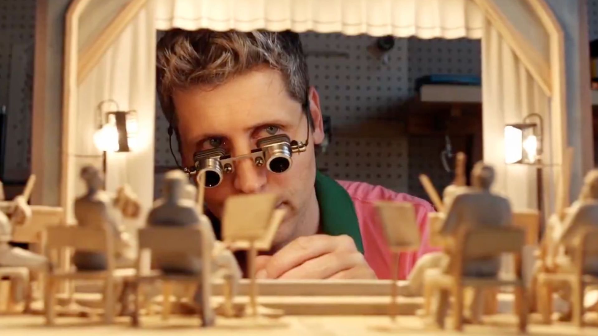 Man with short hair and magnifying glasses working on a detailed miniature orchestra model with figurines holding instruments in a brightly lit workshop.