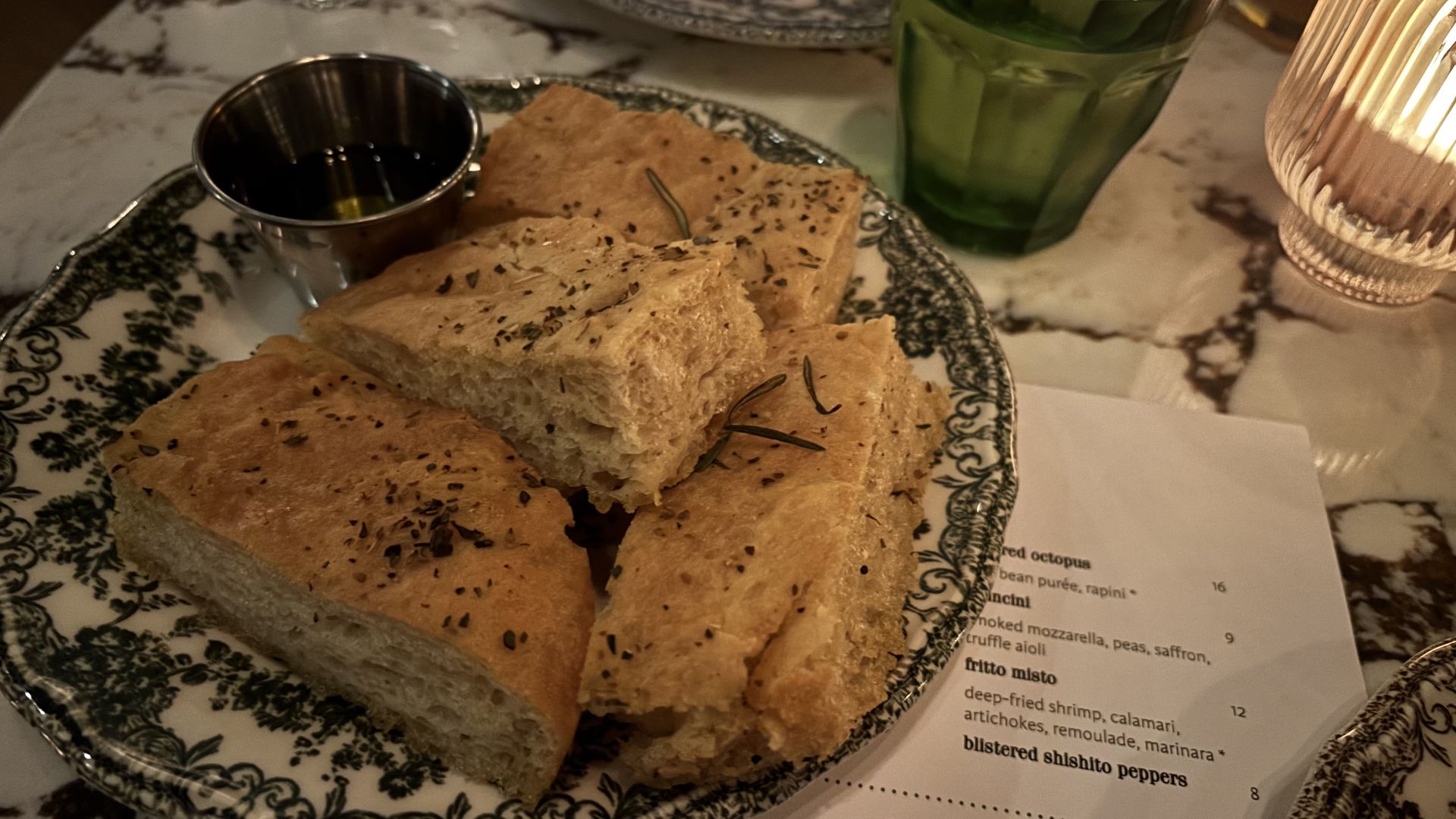 Plate with four rectangular pieces of herb-seasoned focaccia bread, a small metal cup with olive oil, a green glass, and part of a menu on a marble table.