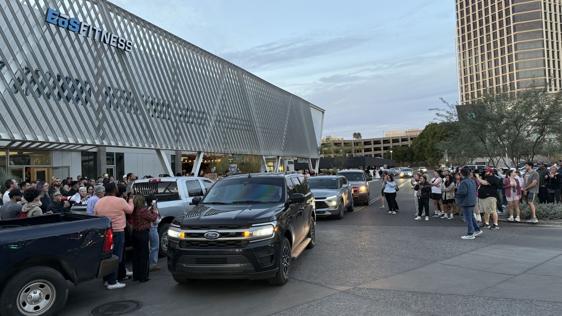 Crowd gathered outside EoS Fitness building during early evening, facing a line of vehicles including black Ford SUV, some people holding cameras and signs.