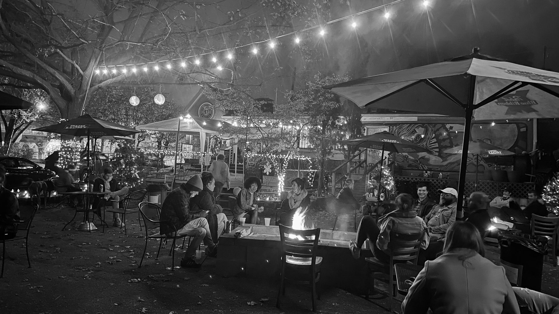 A black-and-white photo of people sitting outdoors around a firepit, at tables, on benches at 97 Estoria