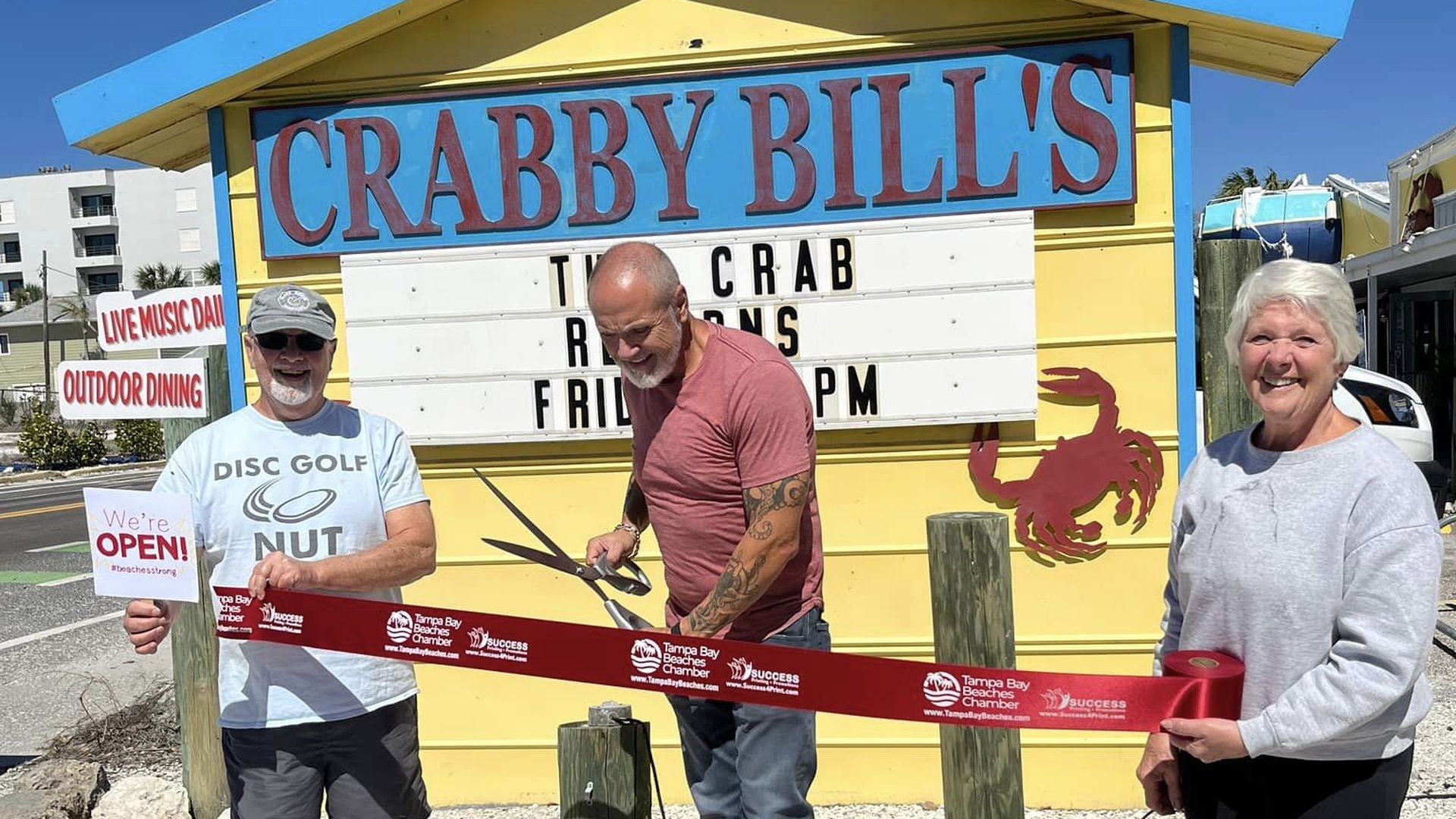 A man with two other people on each side holds a giant pair of scissors about to cut a red ribbon. A yellow-and-blue sign beyond them says, "Crabby Bill's."