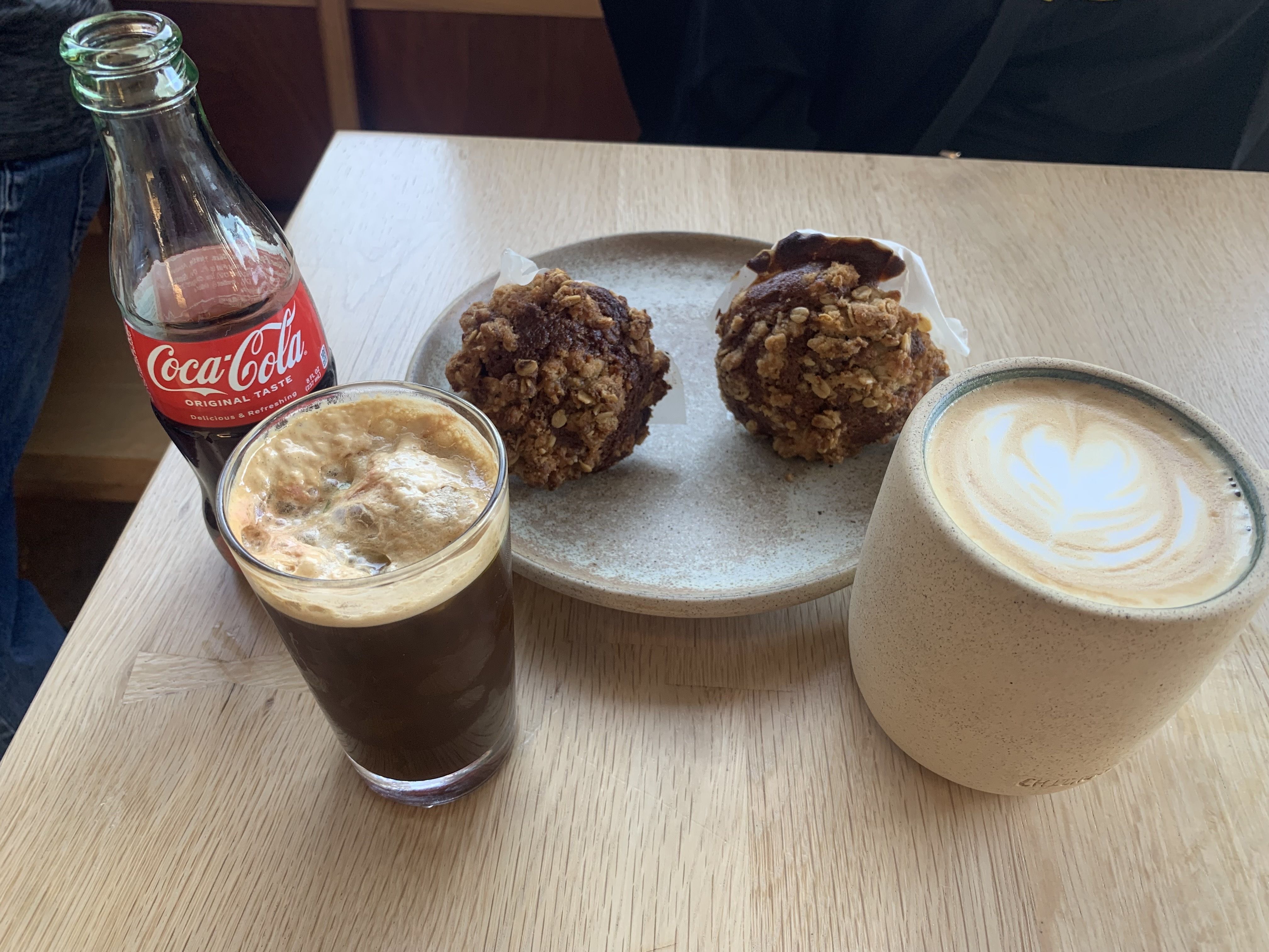 A wooden table with a glass bottle of Coca-Cola, a glass of coffee with foam, two oat-topped muffins on a ceramic plate, and a mug of latte with heart-shaped foam art.