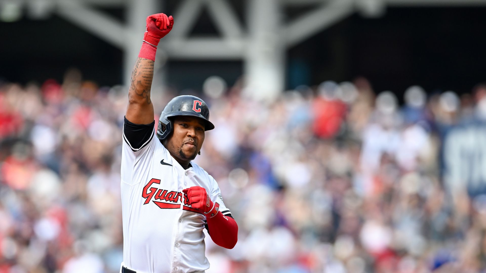 A baseball player in a white Guardians jersey and dark helmet, red gloves, raises his right fist in celebration as a blurred crowd watches from the stands.