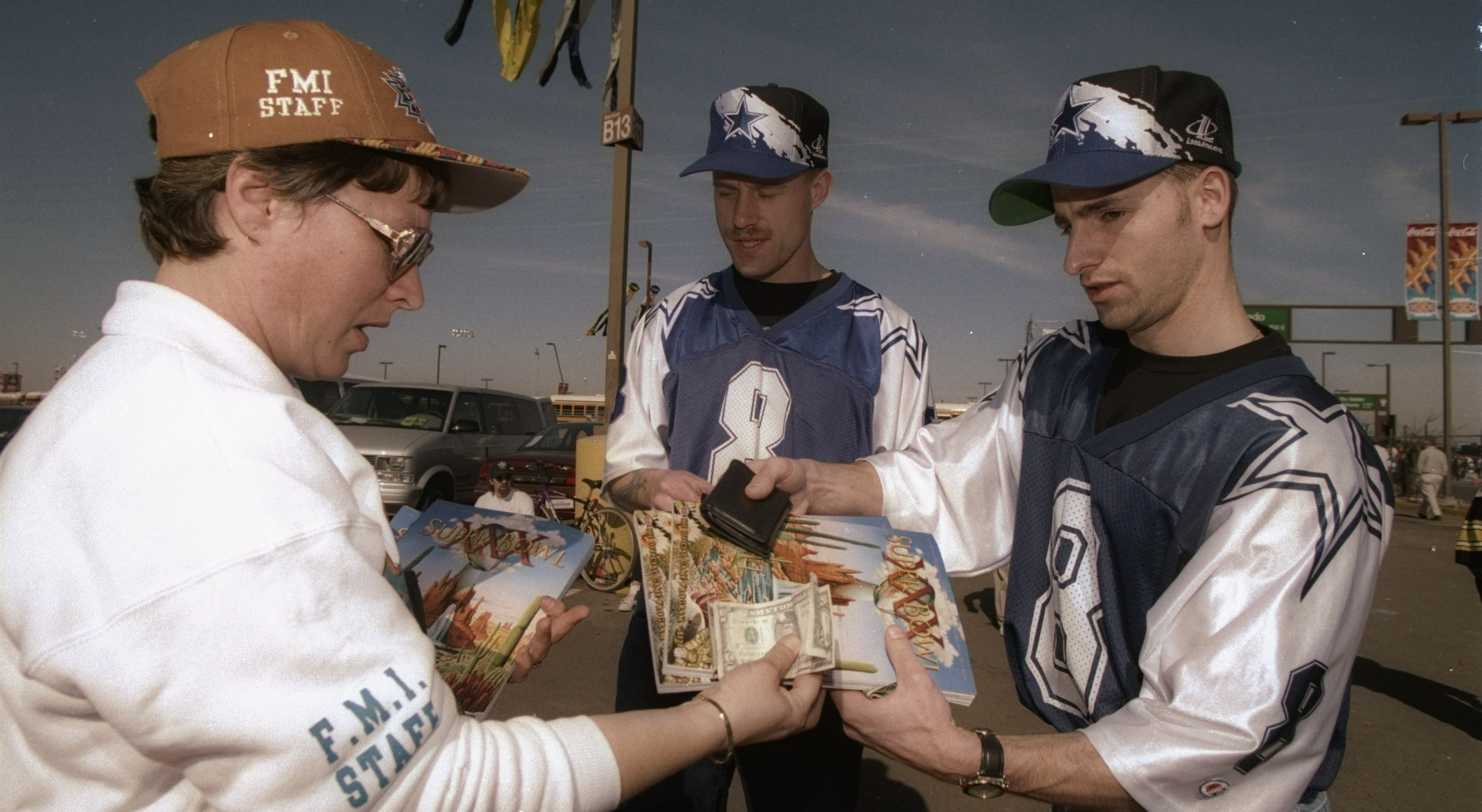 Two men wearing Dallas Cowboys jerseys and hats purchase a game day brochure with cash
