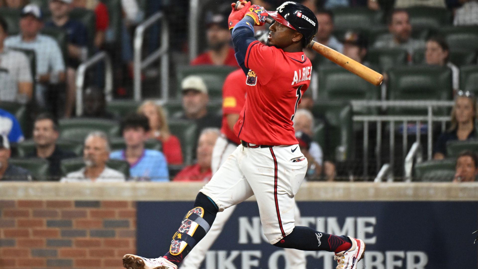 A baseball player wearing a red jersey and white pants with black piping makes contact with a pitch and watches the ball travel toward the fences.