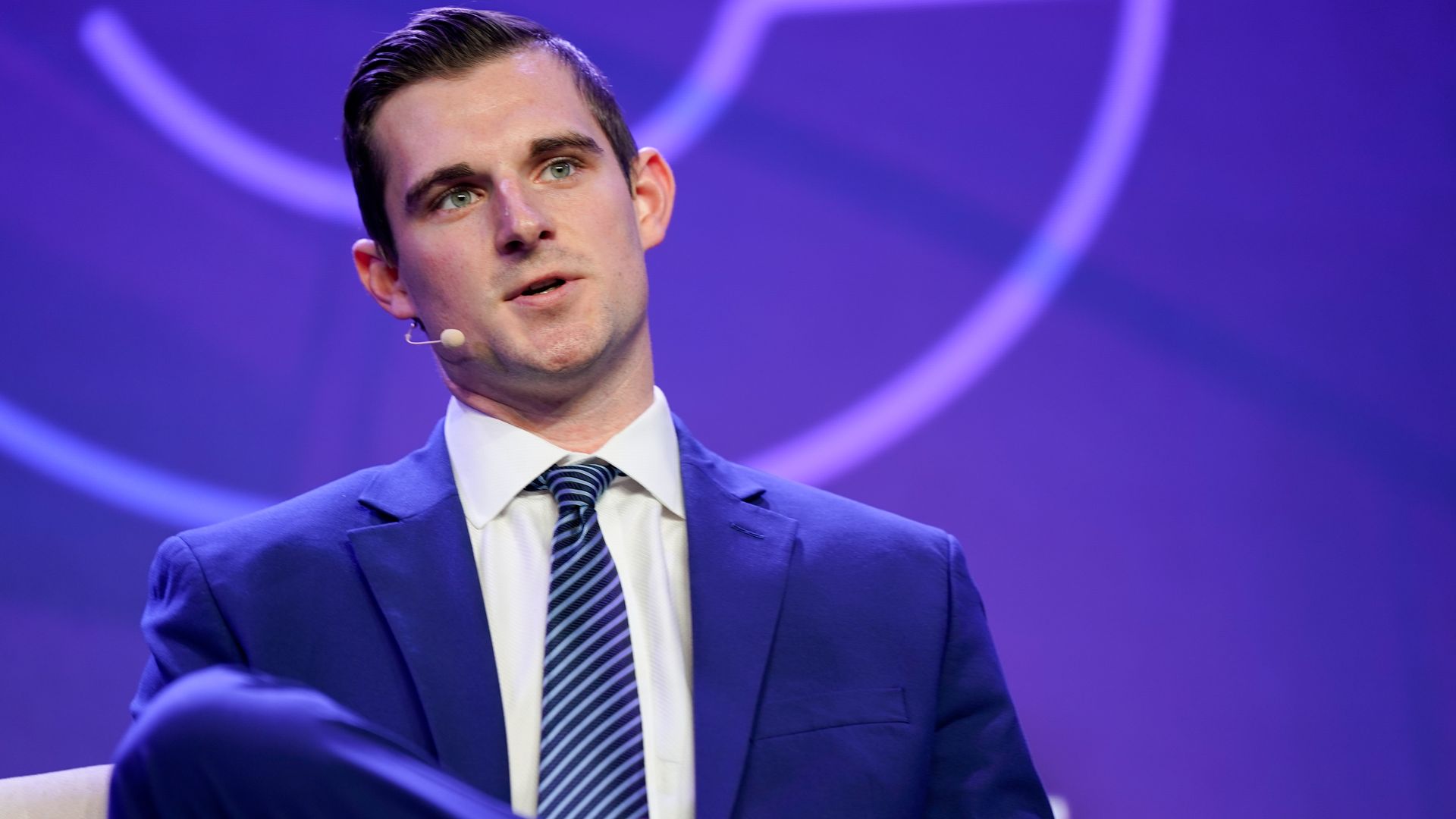 A man speaking in a blue suit, wearing a headset microphone. He's seated, speaking on stage at a large event.