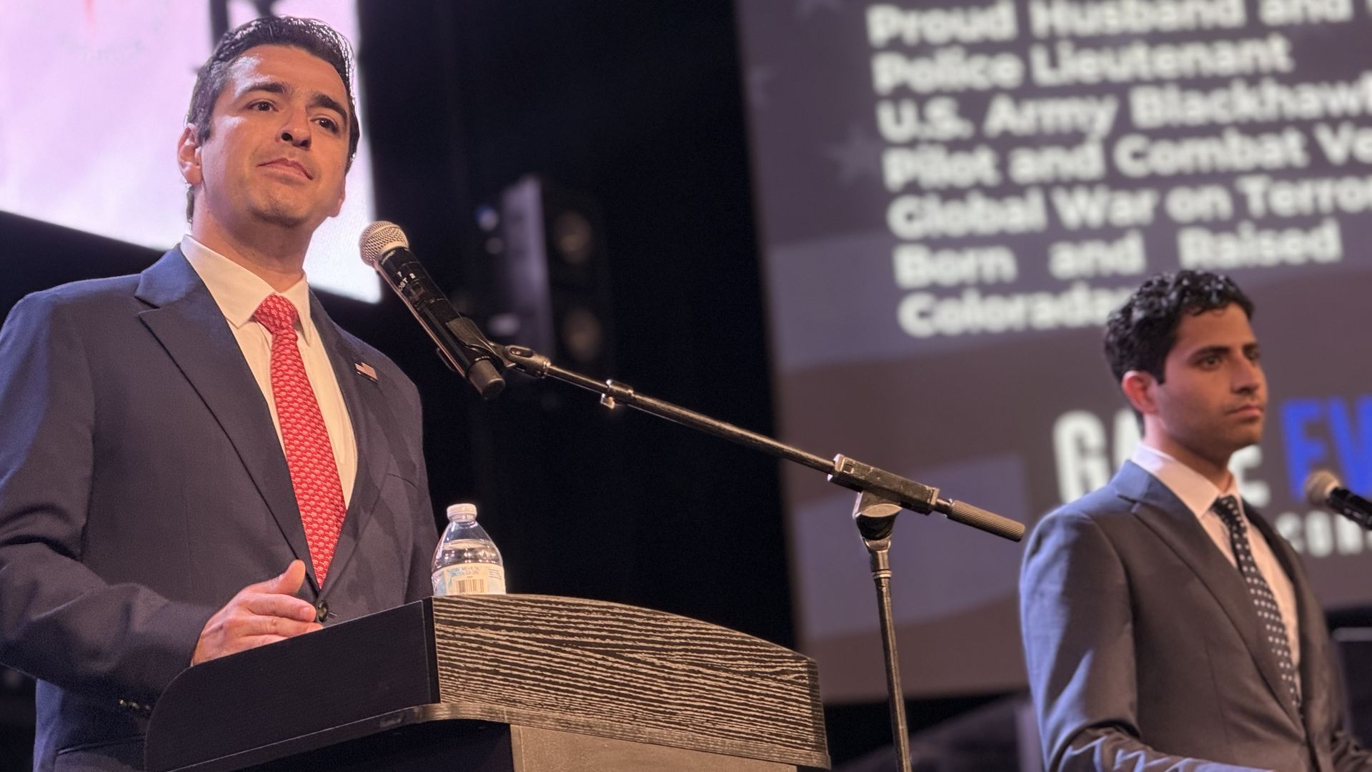 Two men in suits on stage. The left man in a dark blue suit and red tie speaks at a podium with a microphone and water bottle; the right man stands near a blurred screen of text.