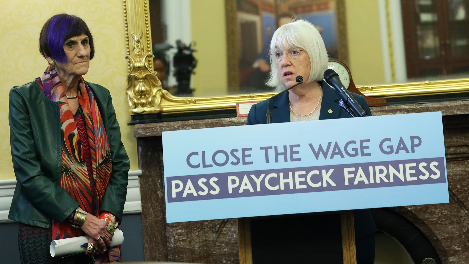 Patty Murray stands at left holding a blue sign that says "Close the Wage Gap: Pass Paycheck Fairness" as she speaks into a microphone, accompanied by another member of Congress.