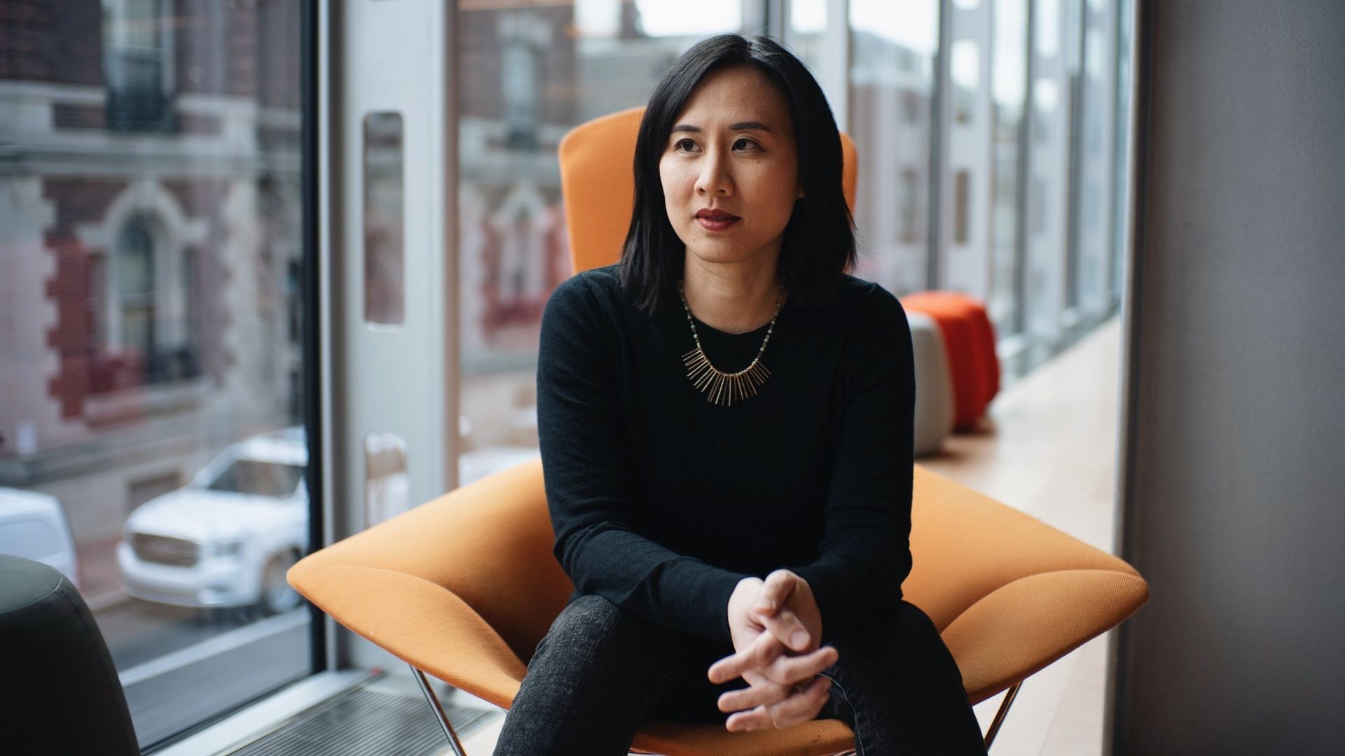 Woman with black hair wearing a black top and necklace sitting on an orange chair near a large window in a modern building.