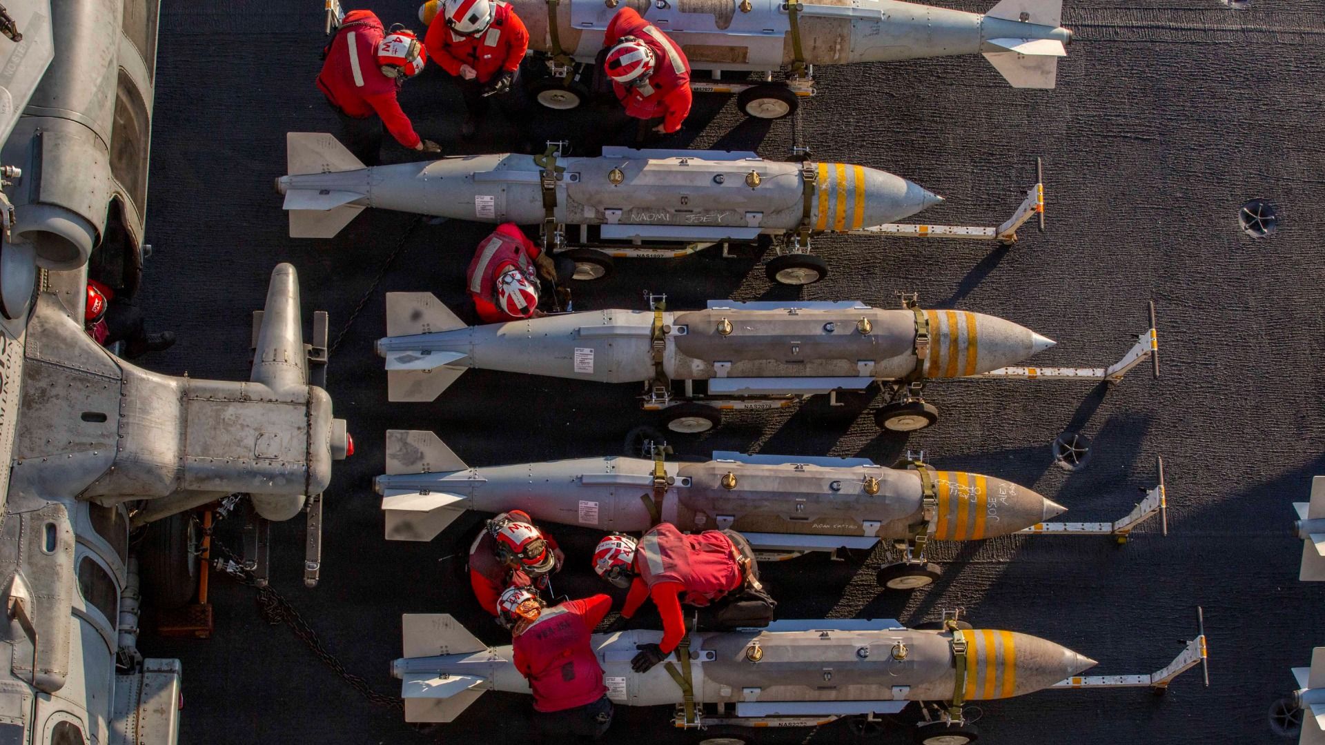 Aerial view of seven people in red outfits working on and around four large gray bombs with yellow stripes, arranged in a row on a dark surface next to part of a gray ship.