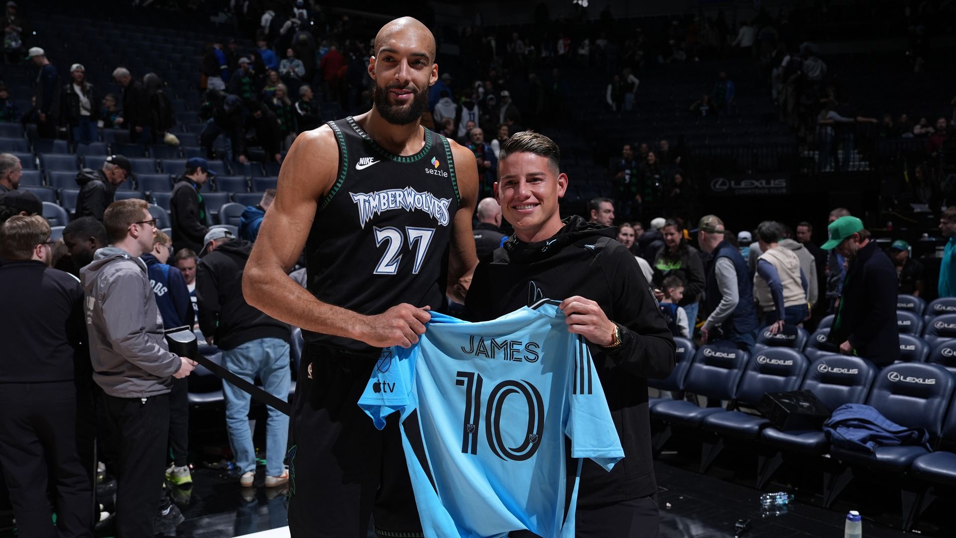 Two men standing on a basketball court; one in a black Timberwolves jersey (#27), the other in a black hoodie holding a blue soccer jersey with name "JAMES" and number 10, with fans in background.