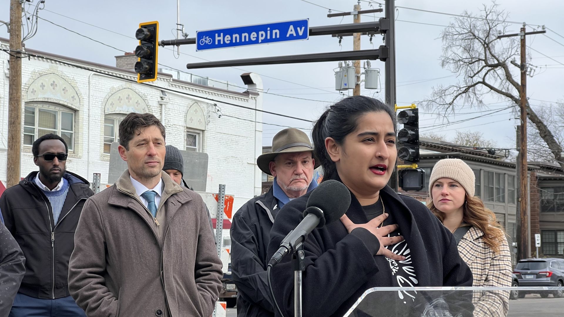 A woman with brown skin stands at a microphone at a news conference under a street sign saying "Hennepin Avenue"