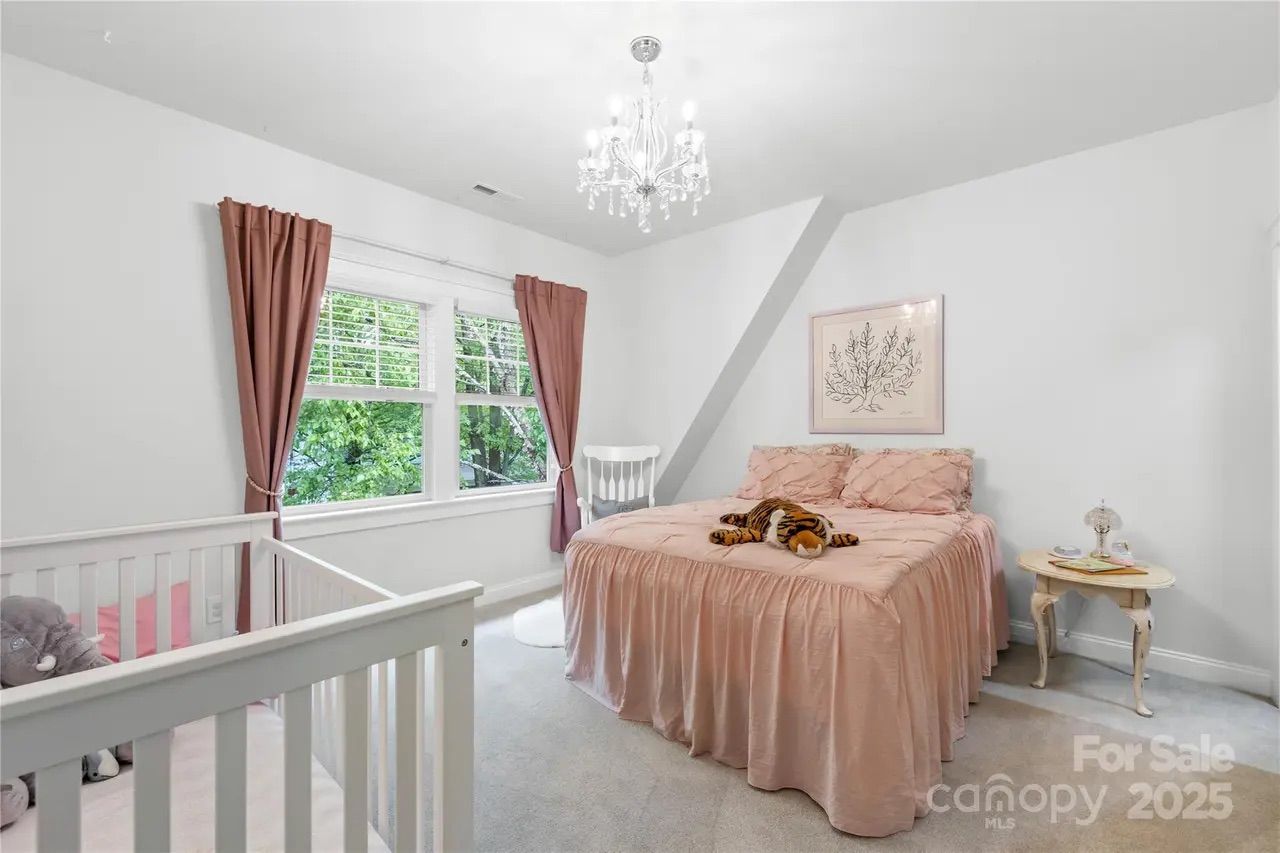 Bright bedroom with white walls, a crib, pink bedspread with stuffed tiger, pink curtains, white rocking chair, small side table, chandelier, and large window showing green trees outside.