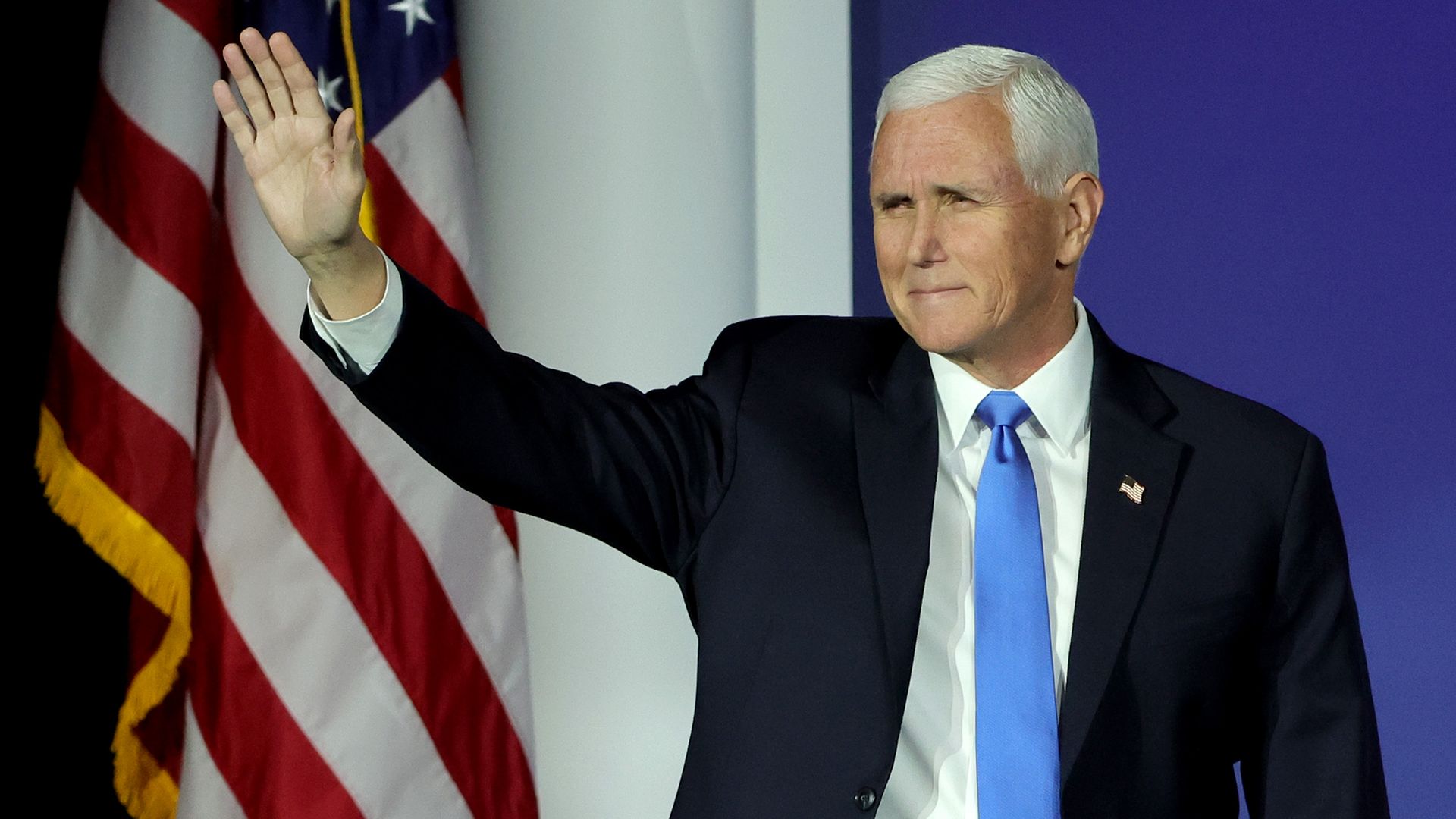 A man in a dark suit and blue tie raises his right hand on stage, with the American flag and a blue background behind him.