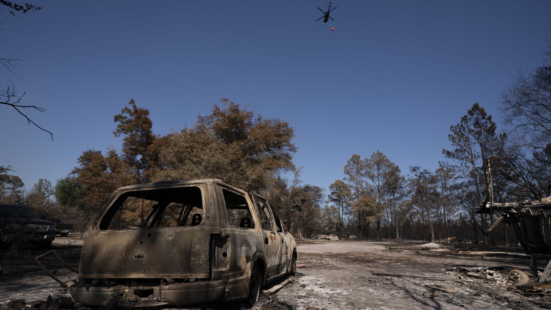 Charred SUV in a scorched lot; a helicopter hovers overhead, dropping a red water bucket as leafless burnt trees and debris fill the background.