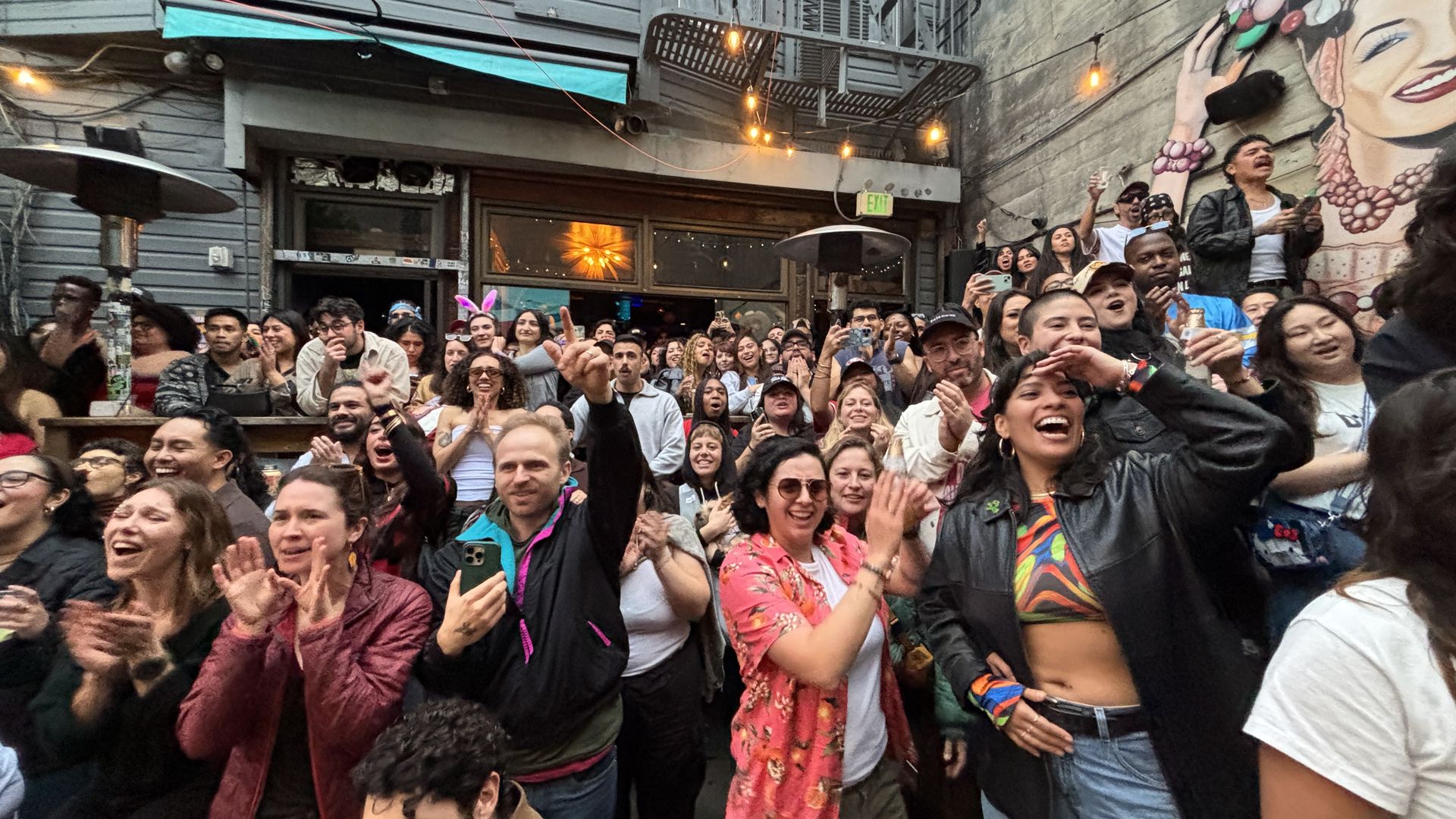 Diverse crowd of happy people clapping and cheering in an outdoor venue with string lights and mural on a concrete wall, some holding phones, one person wearing pink floral shirt, another in black jacket.