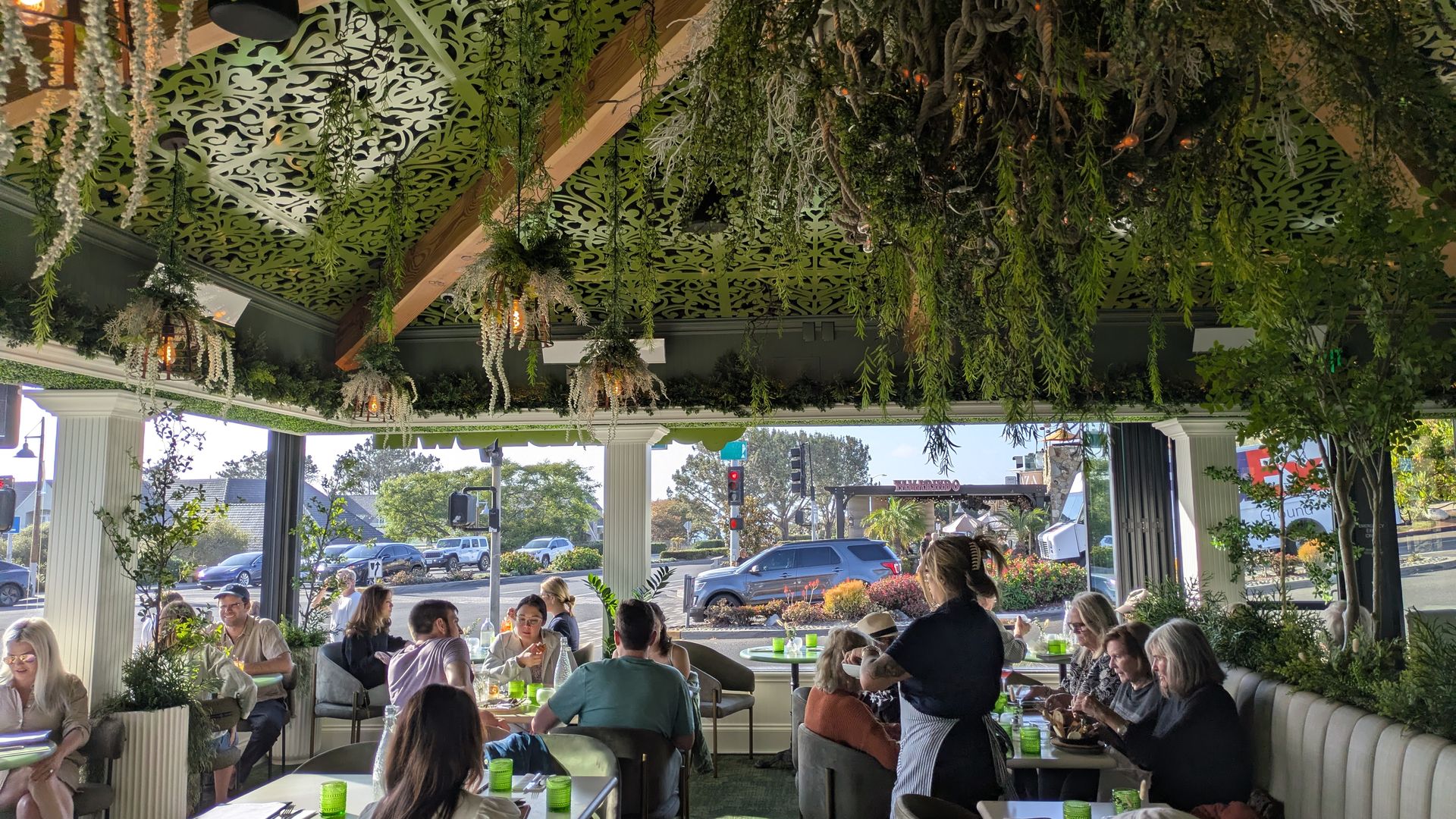 Indoor restaurant with hanging greenery and vines from the ceiling, green patterned panels, and wooden beams. Guests sit at tables with lime-green cups, chatting by large windows.