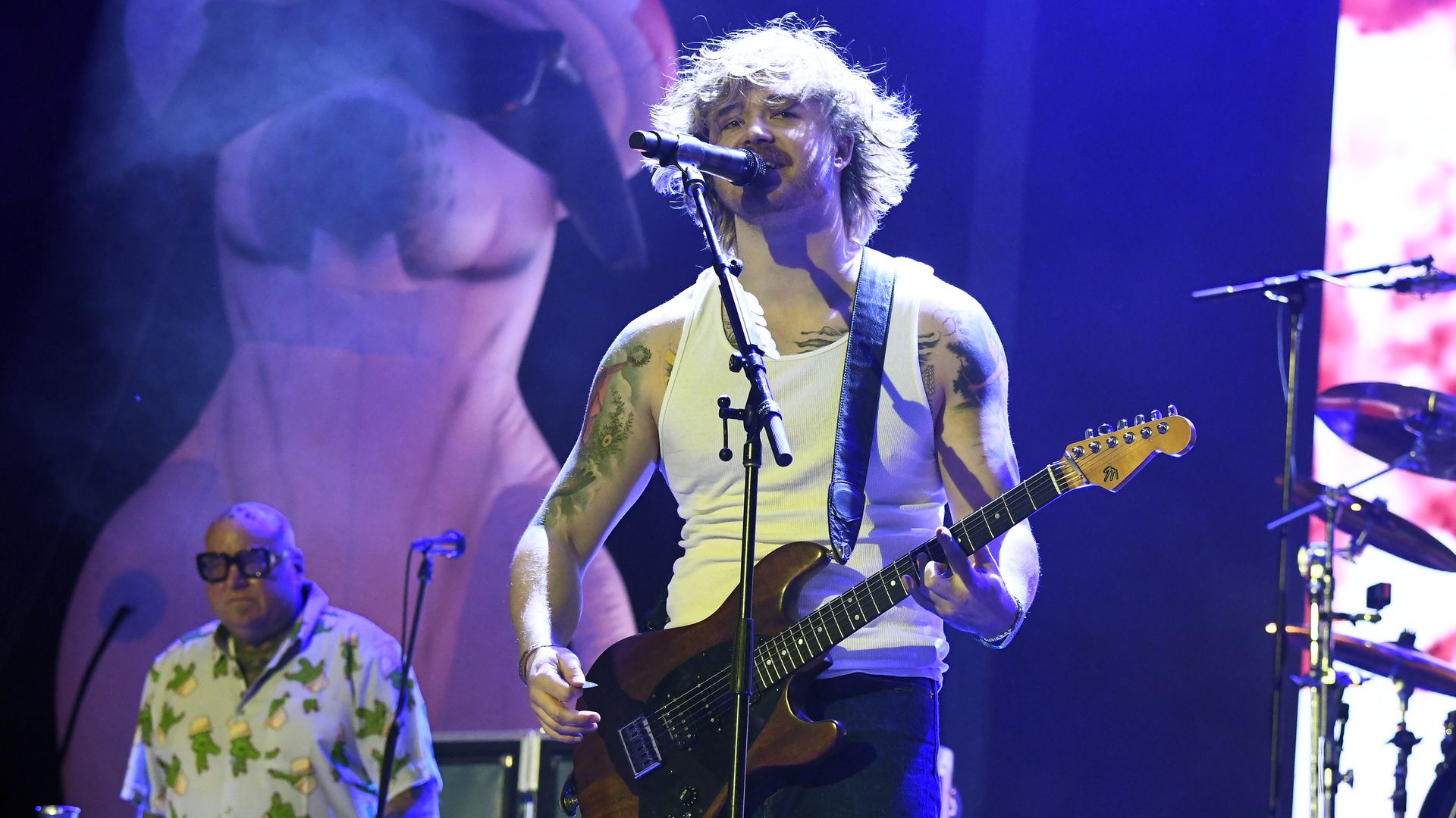Two rockstars — one holding a guitar and singing — perform onstage.