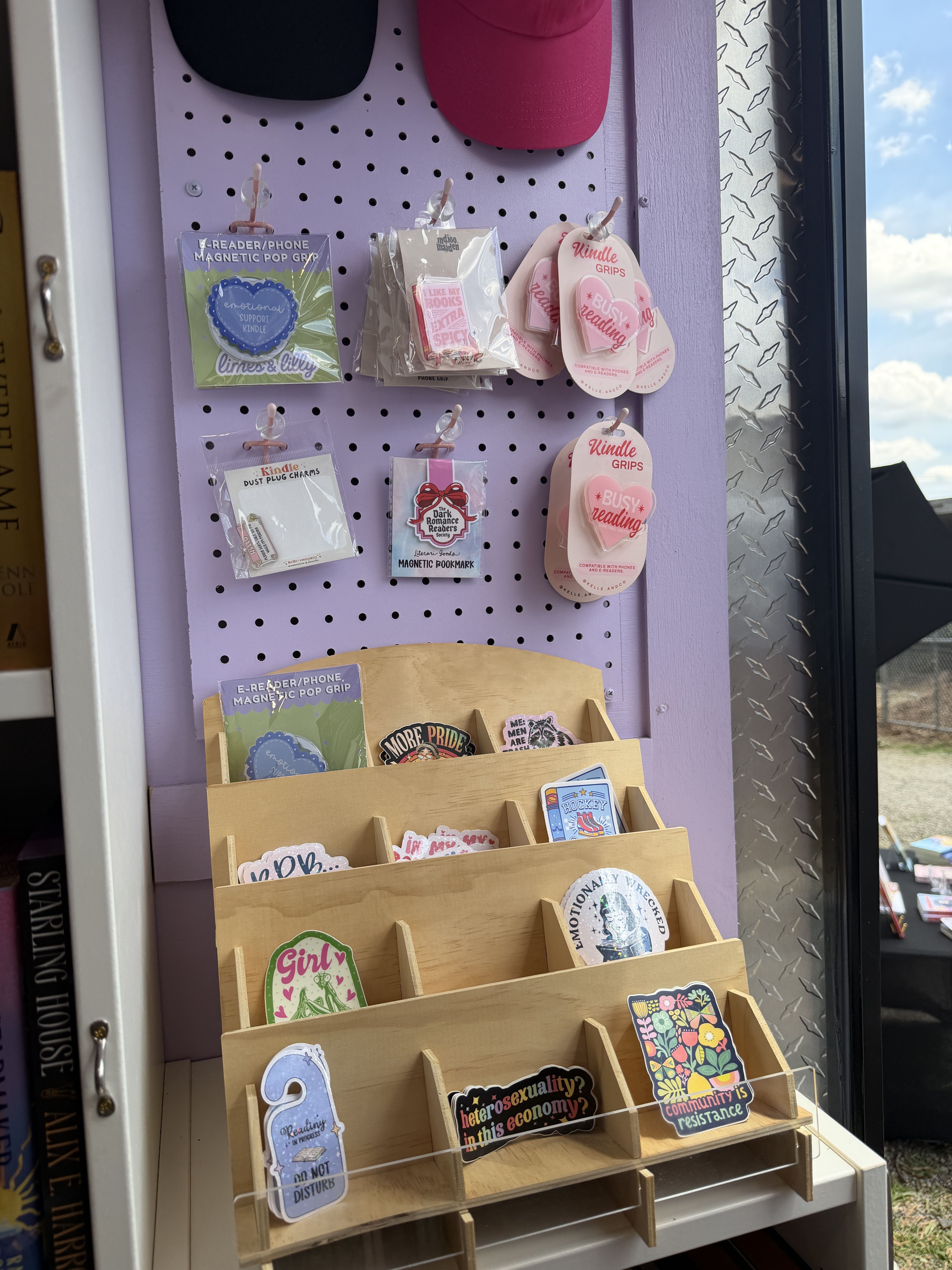 Purple pegboard display with pink hats at the top, showcasing bookish merch: Kindle grips, magnetic pop grips, and colorful stickers/cards arranged on hooks and wooden racks by a window.