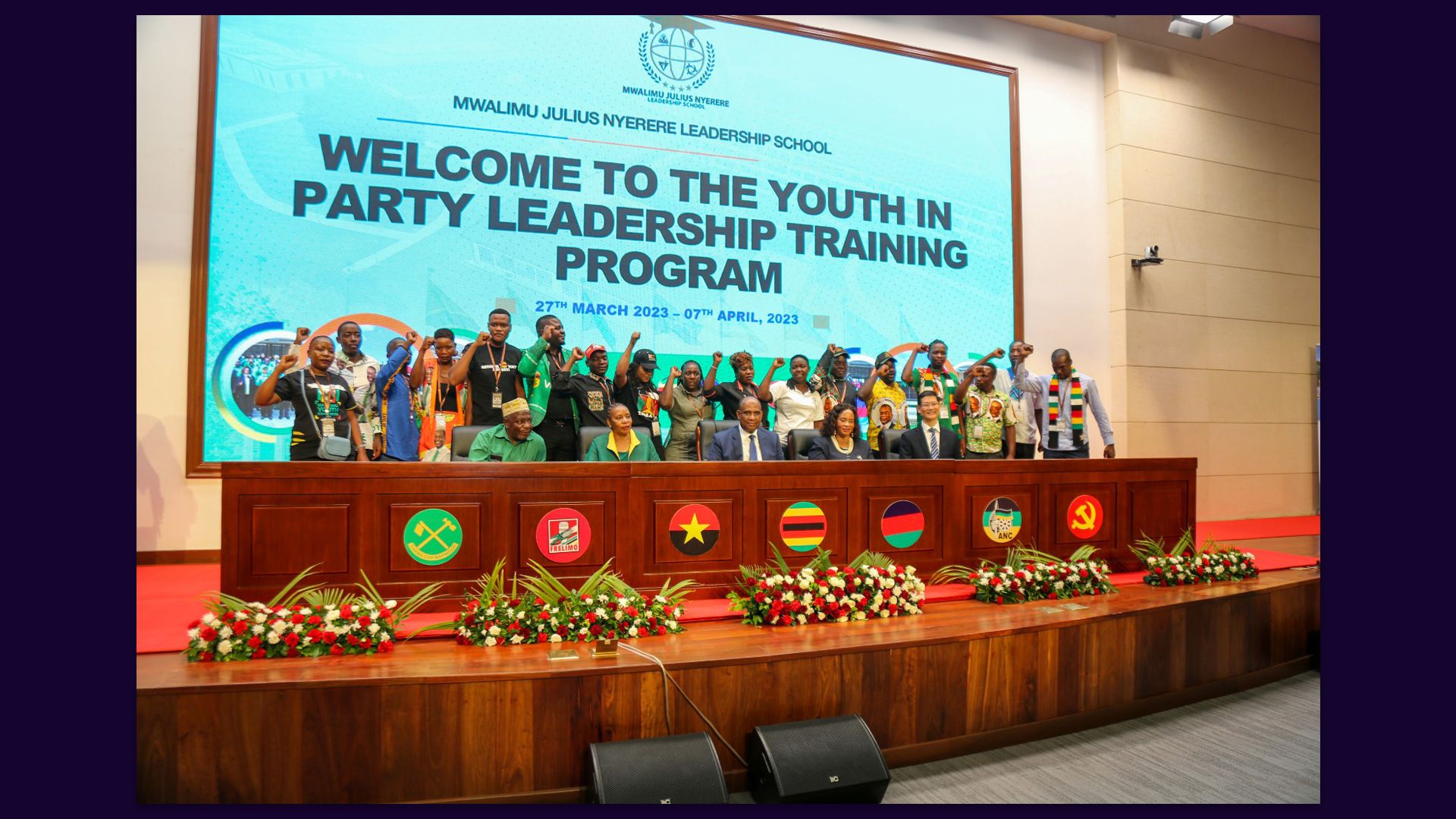 Party leaders sit on stage. Behind them are participants with their fists raised, and a presentation slide that reads, “Welcome to the youth in party leadership training program; March 27, 2023 to April 7, 2023.”