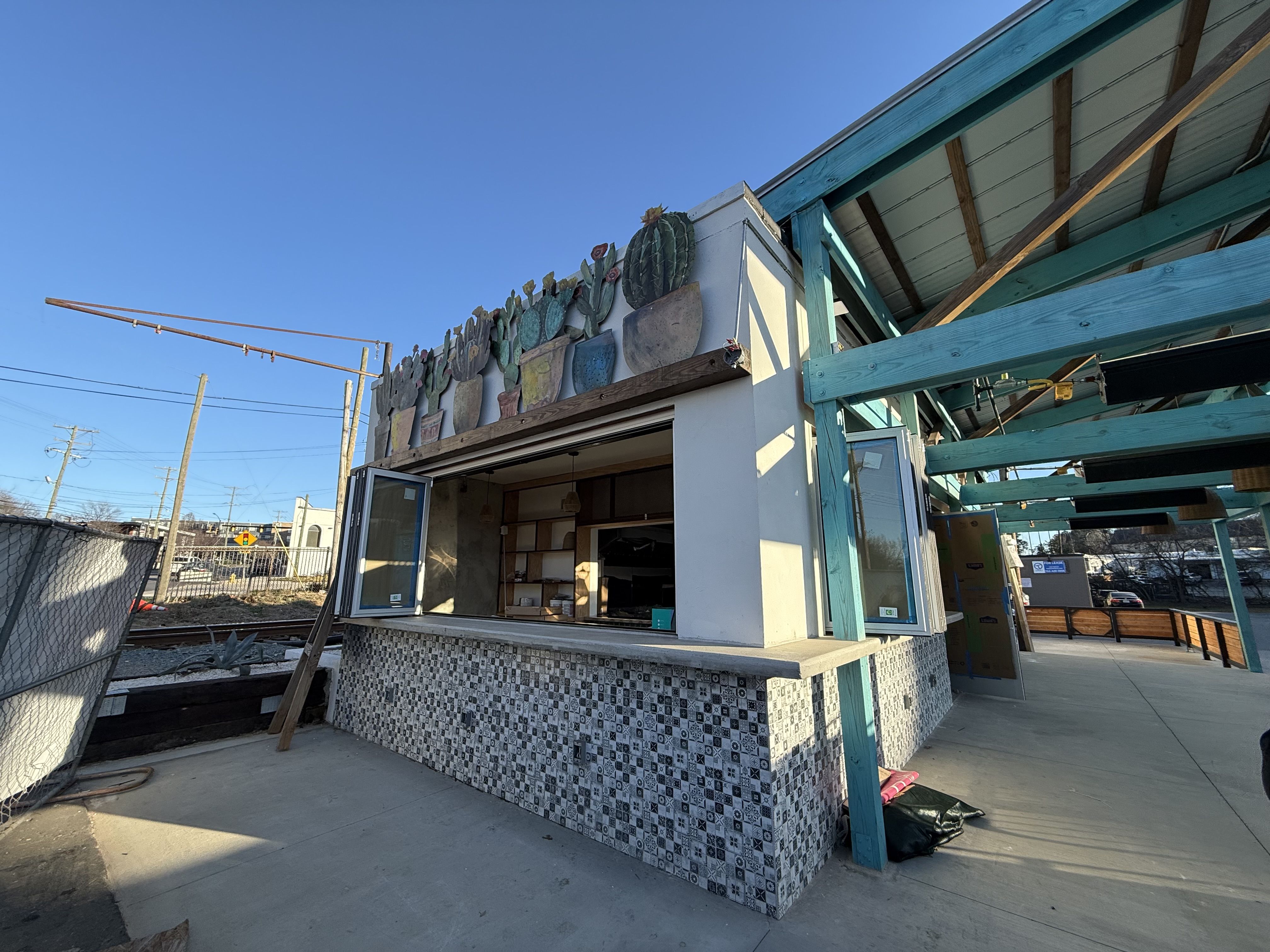 Outdoor food stand with open windows, decorated with colorful ceramic cactus pots on top. The counter is tiled in black and white patterns. Blue wooden beams support an overhead roof.