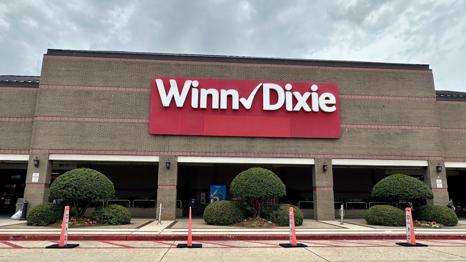 Winn-Dixie store facade: beige brick exterior with a large red sign reading "Winn-Dixie" in white letters; foreground shows trimmed bushes, orange safety cones, and a cloudy sky above.
