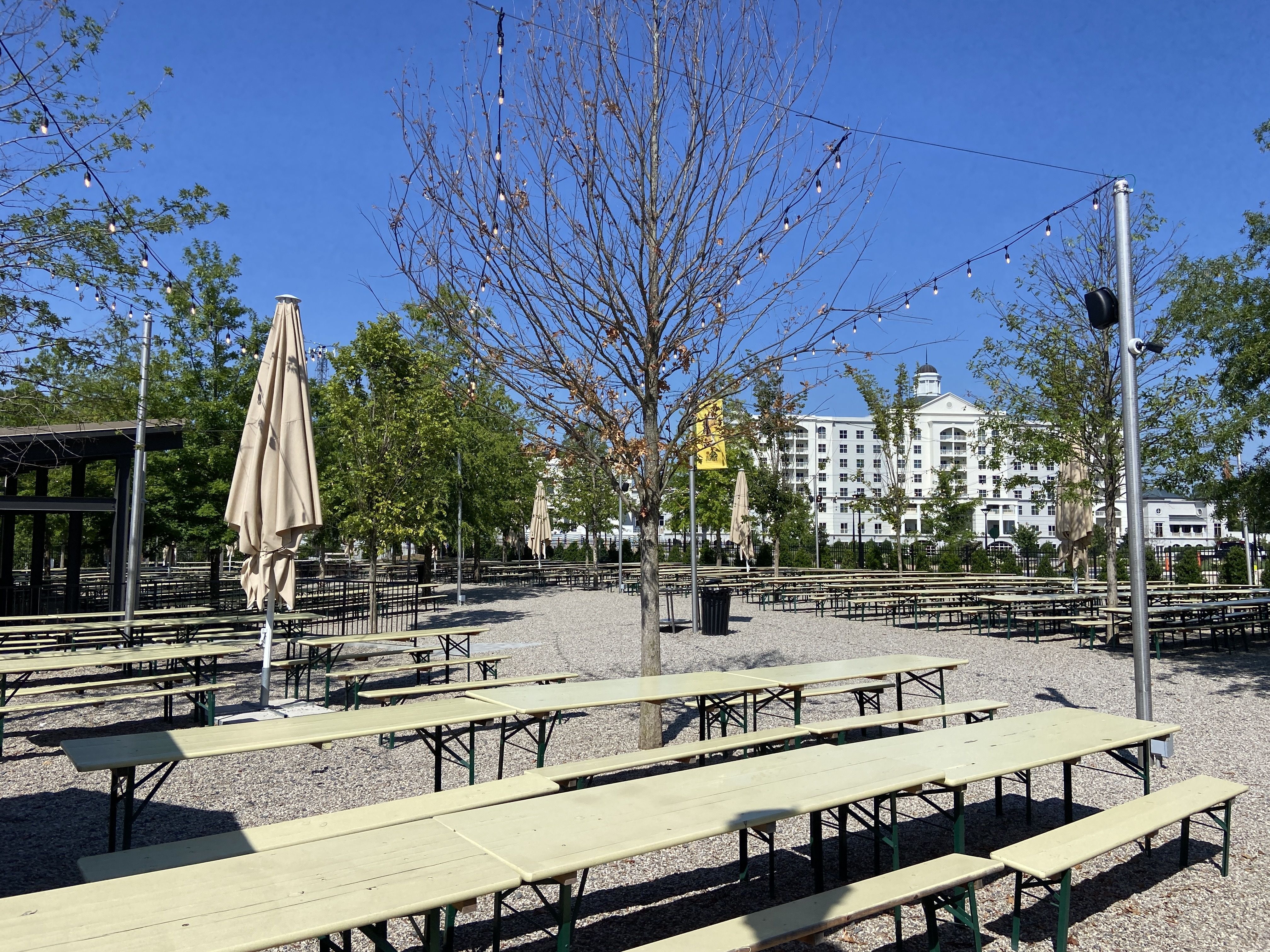 OMB's Ballantyne beer garden with long beige picnic tables, closed beige umbrellas, string lights hanging above, leafless and green trees, and the Ballantyne Hotel in the background under blue sky.