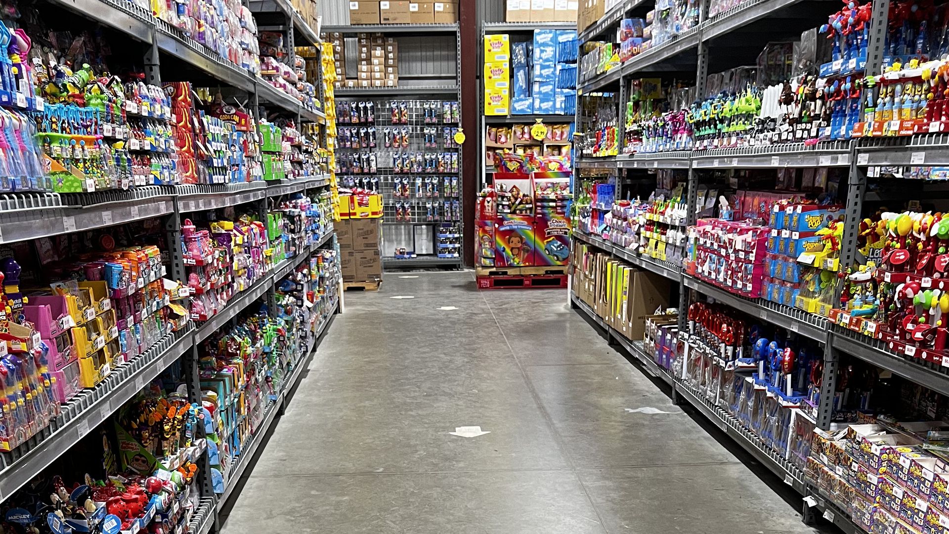 A young child in a colorful jacket pushes a blue plastic shopping cart down a toy aisle in a store, surrounded by shelves filled with various colorful toys.