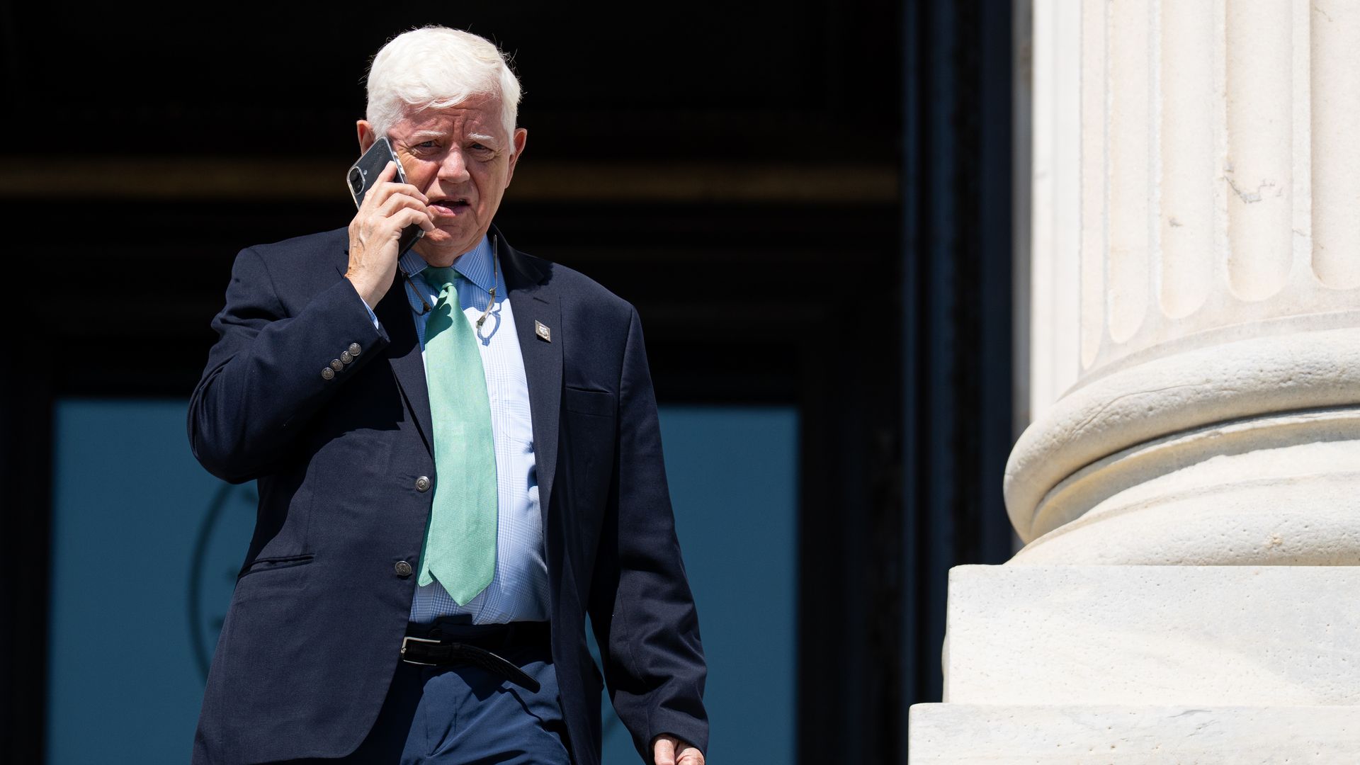 A man with white hair, wearing a navy suit, light blue shirt, and green tie, speaks on a smartphone while standing beside a large stone column.