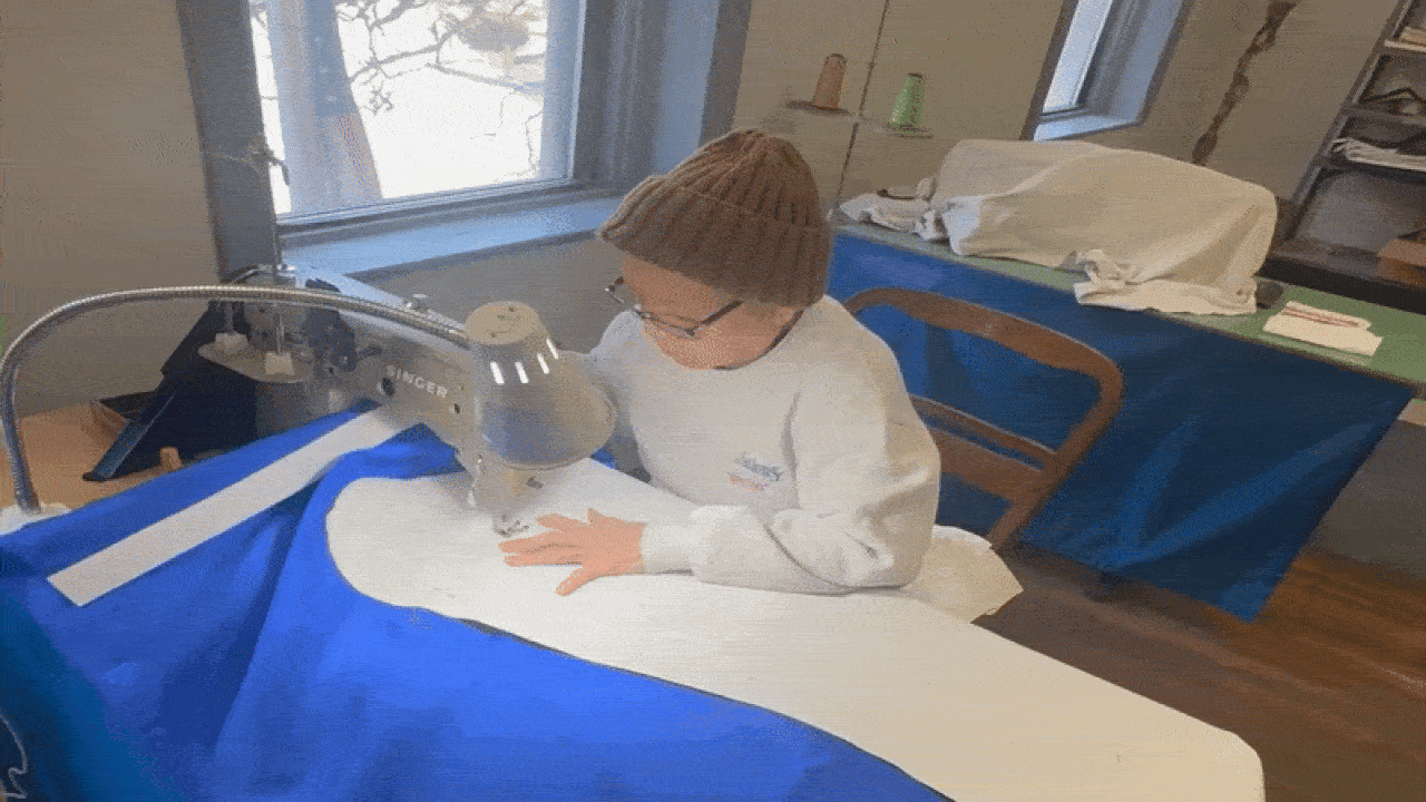 Person wearing a brown knit hat and white sweatshirt sewing large white fabric piece onto blue fabric using a Singer sewing machine in a well-lit room with wooden floors and a window.