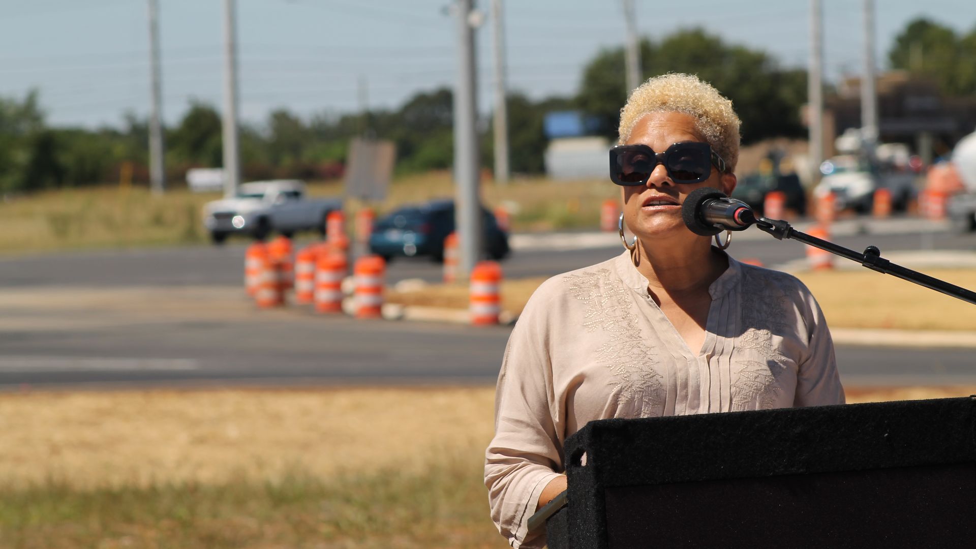 Woman with short blonde curly hair and large black sunglasses speaking at an outdoor podium with a microphone, near a road with orange traffic barrels in the background.
