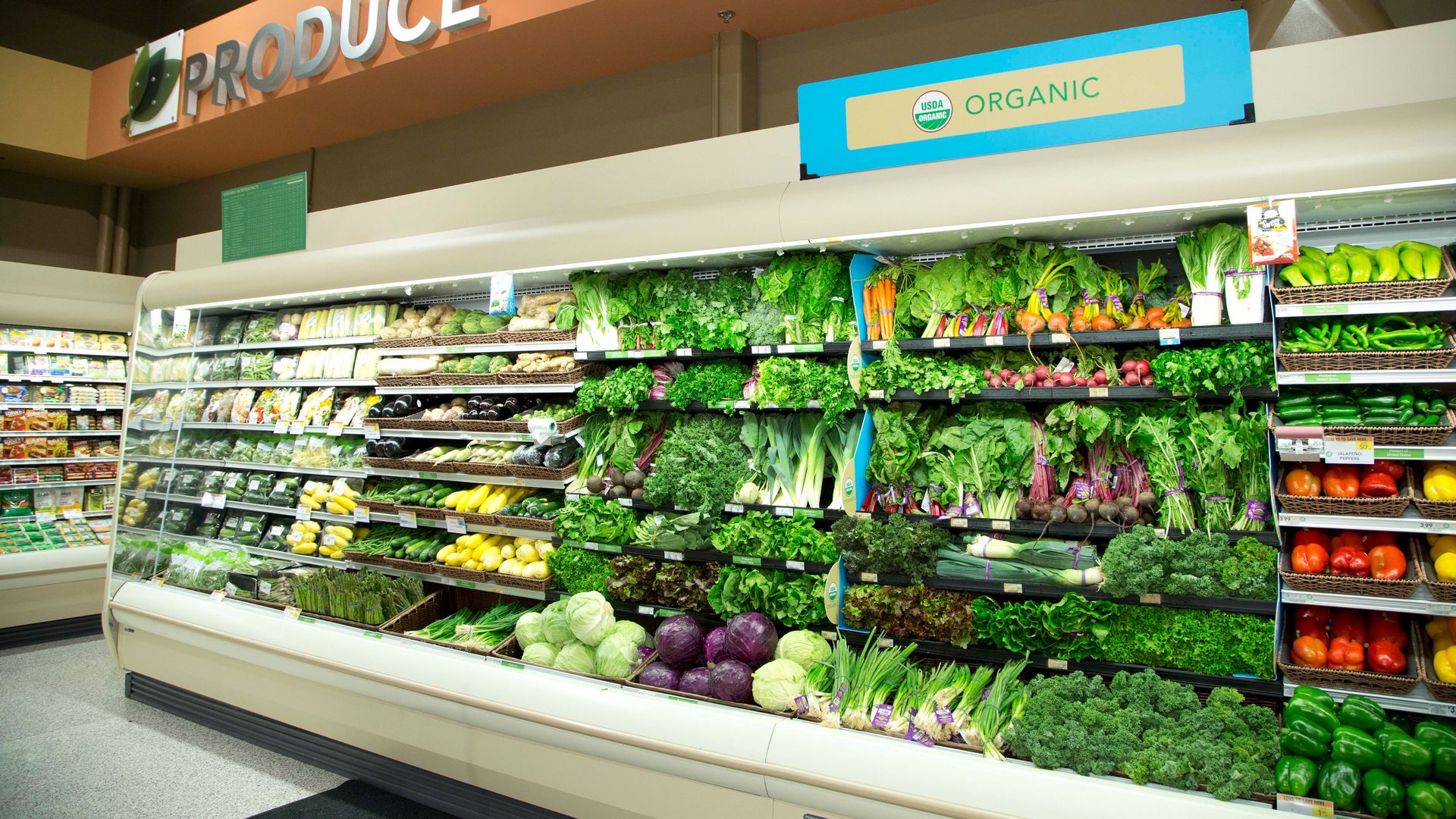 Organic produce section in a grocery store with a variety of vegetables including leafy greens, carrots, cabbage, peppers, and zucchini under an "Organic" USDA label and a large "Produce" sign.