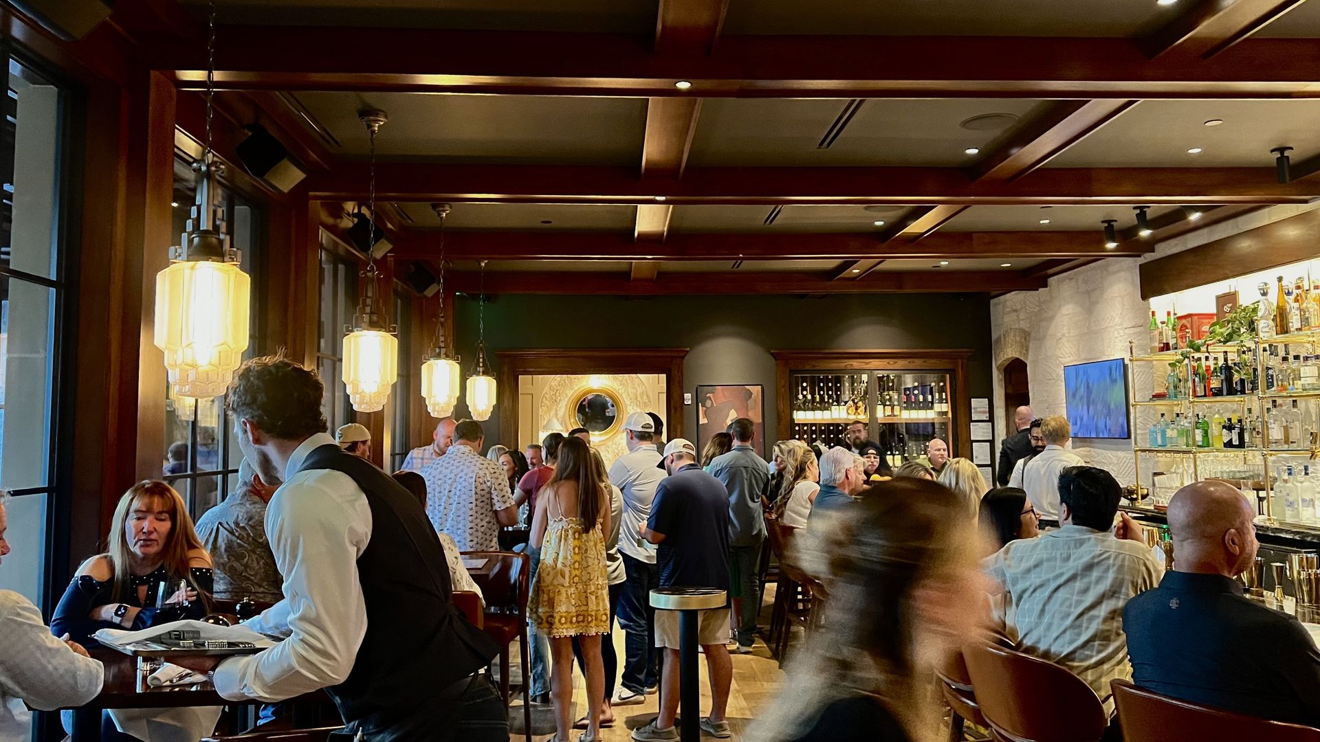 Busy upscale bar and dining area with warm pendant lights, dark wood beams, and a long backlit liquor shelf on the right. Numerous patrons sit and mingle, while a waiter in a vest serves at tables.