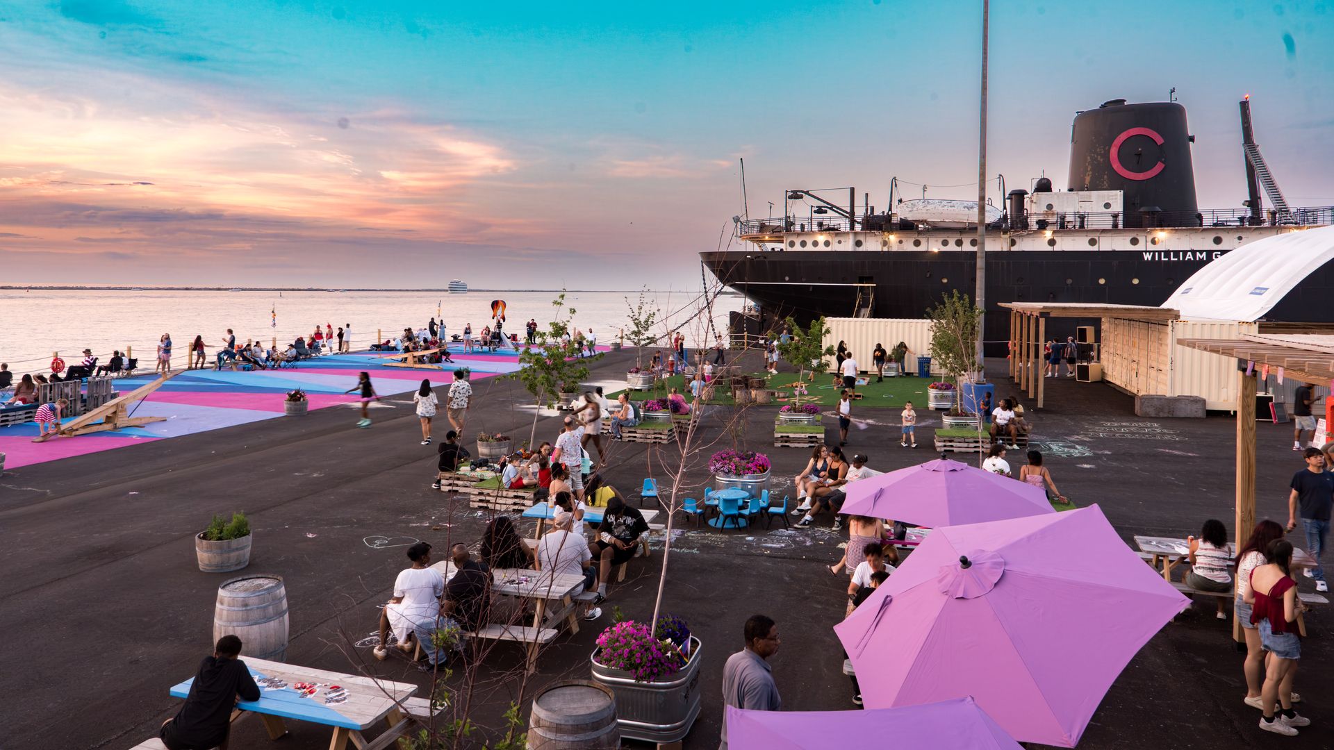 Sunset over a harbor: a large dark ship with a pink C on its funnel docked beside a lively crowd on colorful mats, purple umbrellas, and blue water.