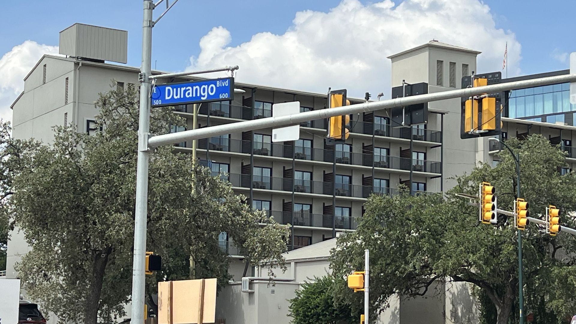 Urban intersection featuring a blue Durango Blvd street sign on a gray pole, modern apartment building, yellow traffic lights, blue sky with clouds, and construction barriers in foreground.