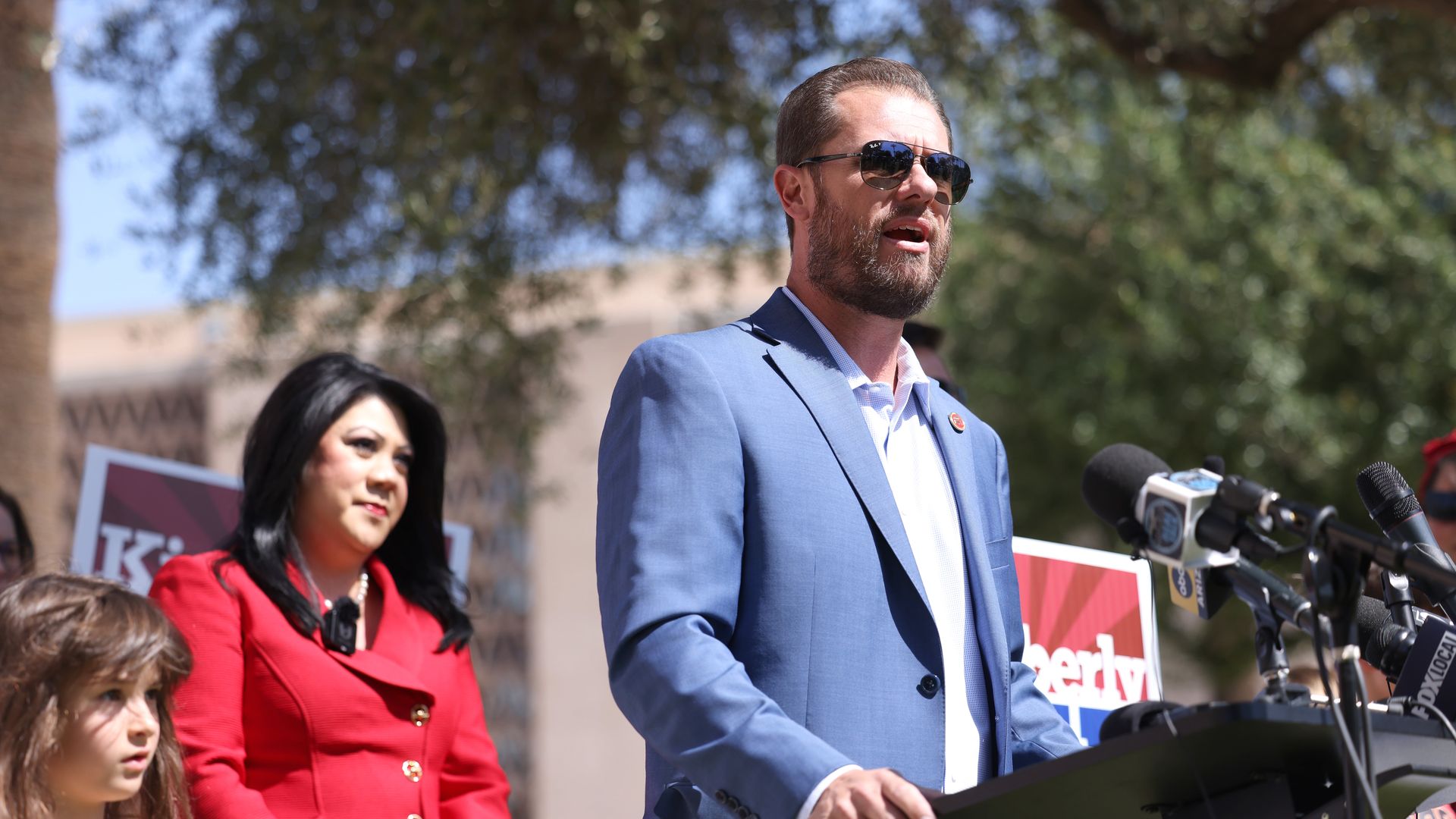 Jake Hoffman in a blue suit and sunglasses speaks at a podium with microphones during an outdoor press conference; Kimberly Yee in red and a child stand nearby, with trees behind.