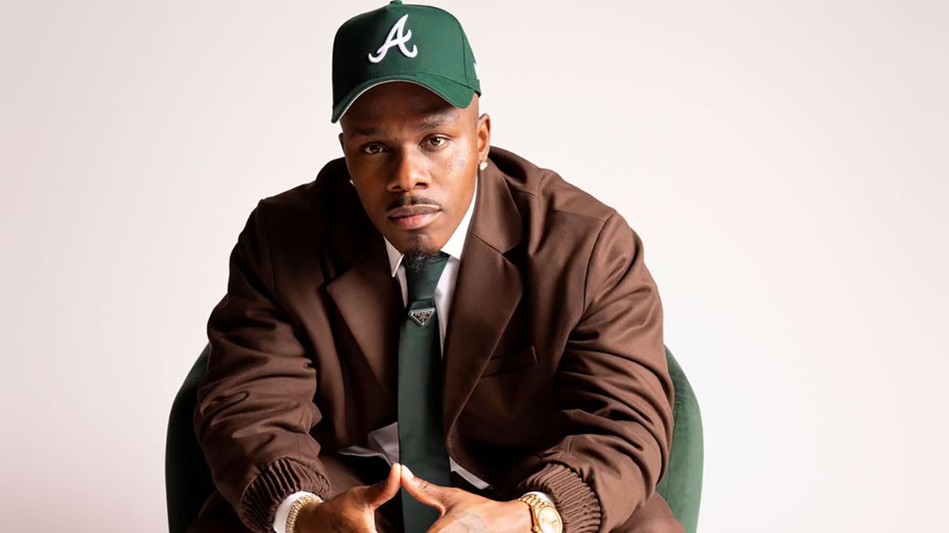 Dababy. Charlotte restaurant. Portrait of a man in a brown suit and a green cap, sitting on a green chair. He wears a green tie, white sneakers, and a gold watch, with hands clasped and a serious expression.