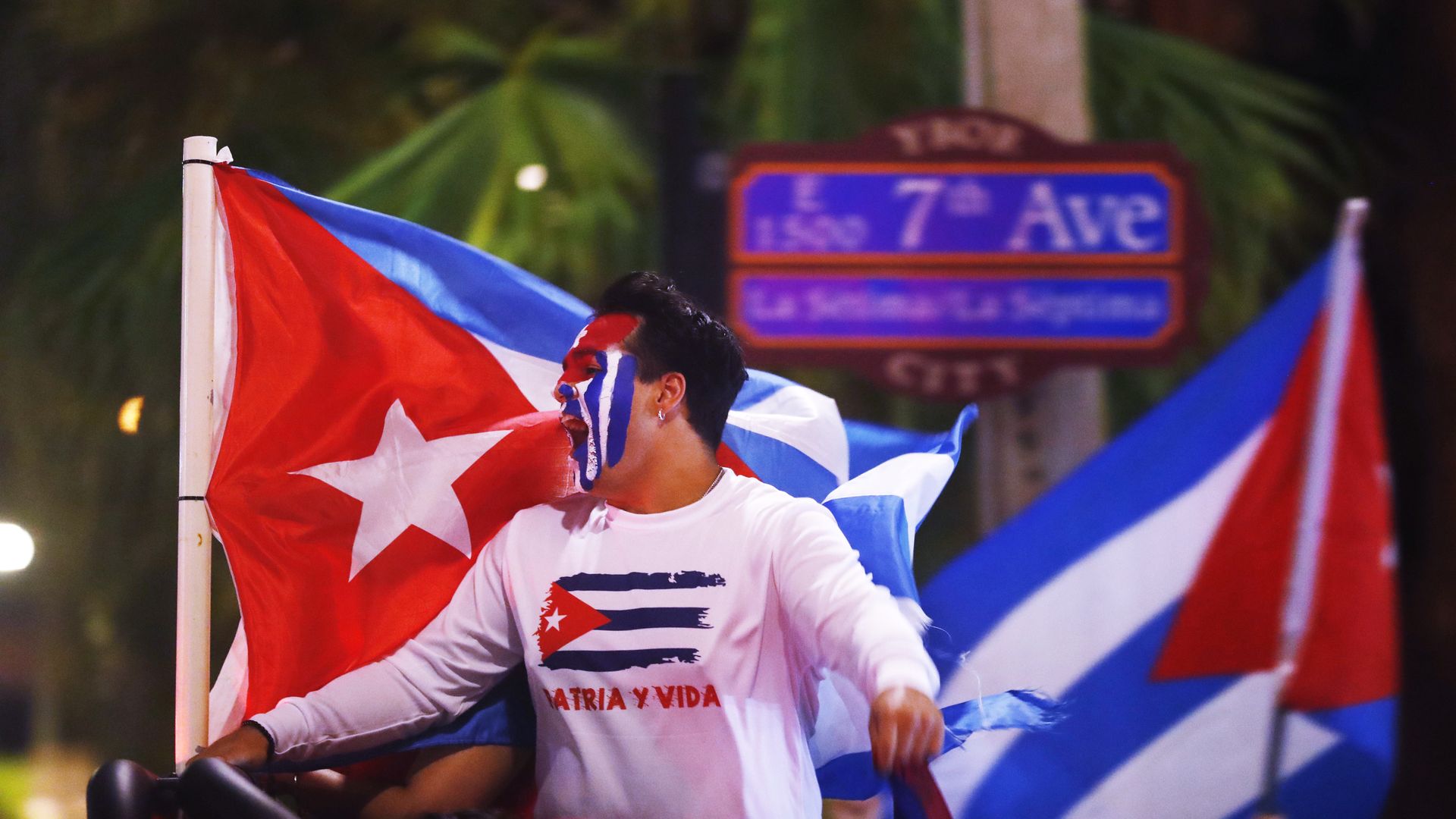 a man with the cuban flag painted on his face holds a cuban flag in front of a sign for 7th ave in ybor